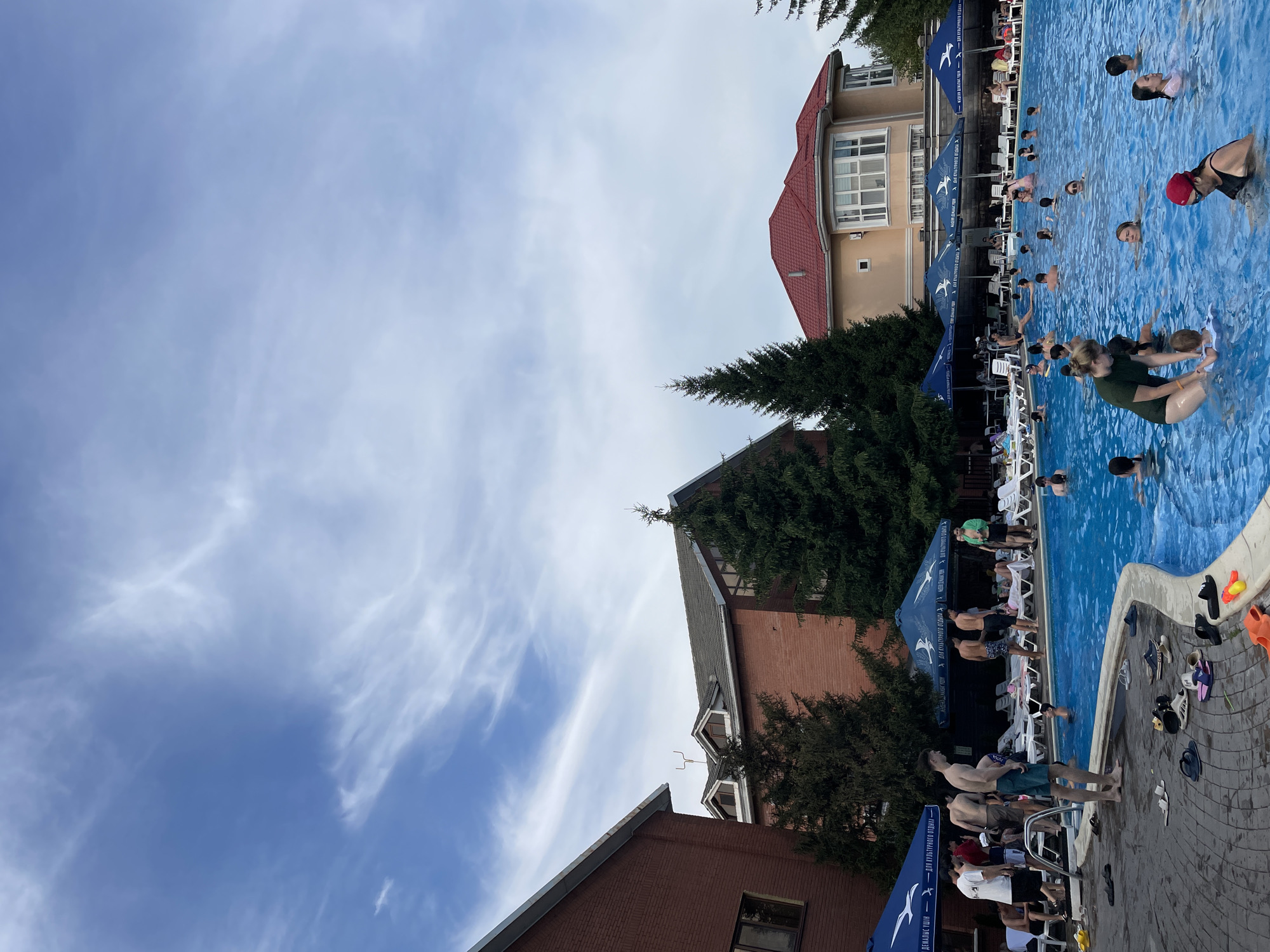 The image depicts an outdoor swimming pool area at what appears to be a resort or recreational facility. The pool is filled with blue water and many people are swimming and wading. Several individuals are standing around the edge of the pool, some are in swimsuits. There are lounge chairs and umbrellas surrounding the pool area. In the background, there are buildings with red tiled roofs and brick exteriors, as well as mature evergreen trees. The sky is partly cloudy with wispy white clouds against a blue background. While the image quality and perspective make it impossible to definitively identify the location within Almaty, Kazakhstan, the architecture and presence of an outdoor pool suggest it could be a hotel, a water park, or a private club.