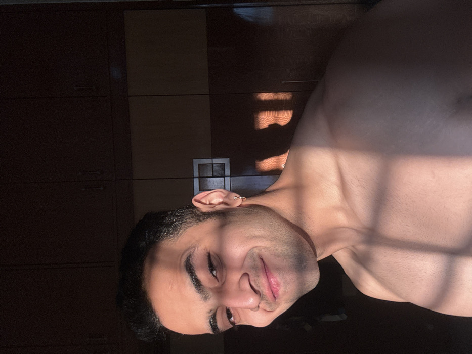 A shirtless man is lying down, illuminated by strong sunlight filtering through what appears to be a window or blinds. The light casts shadows on his upper chest and neck. He is gazing at the camera with a subtle smile. He has short, dark hair and what appears to be a small ring piercing in his left ear. Behind him is a dark brown closet or wardrobe.