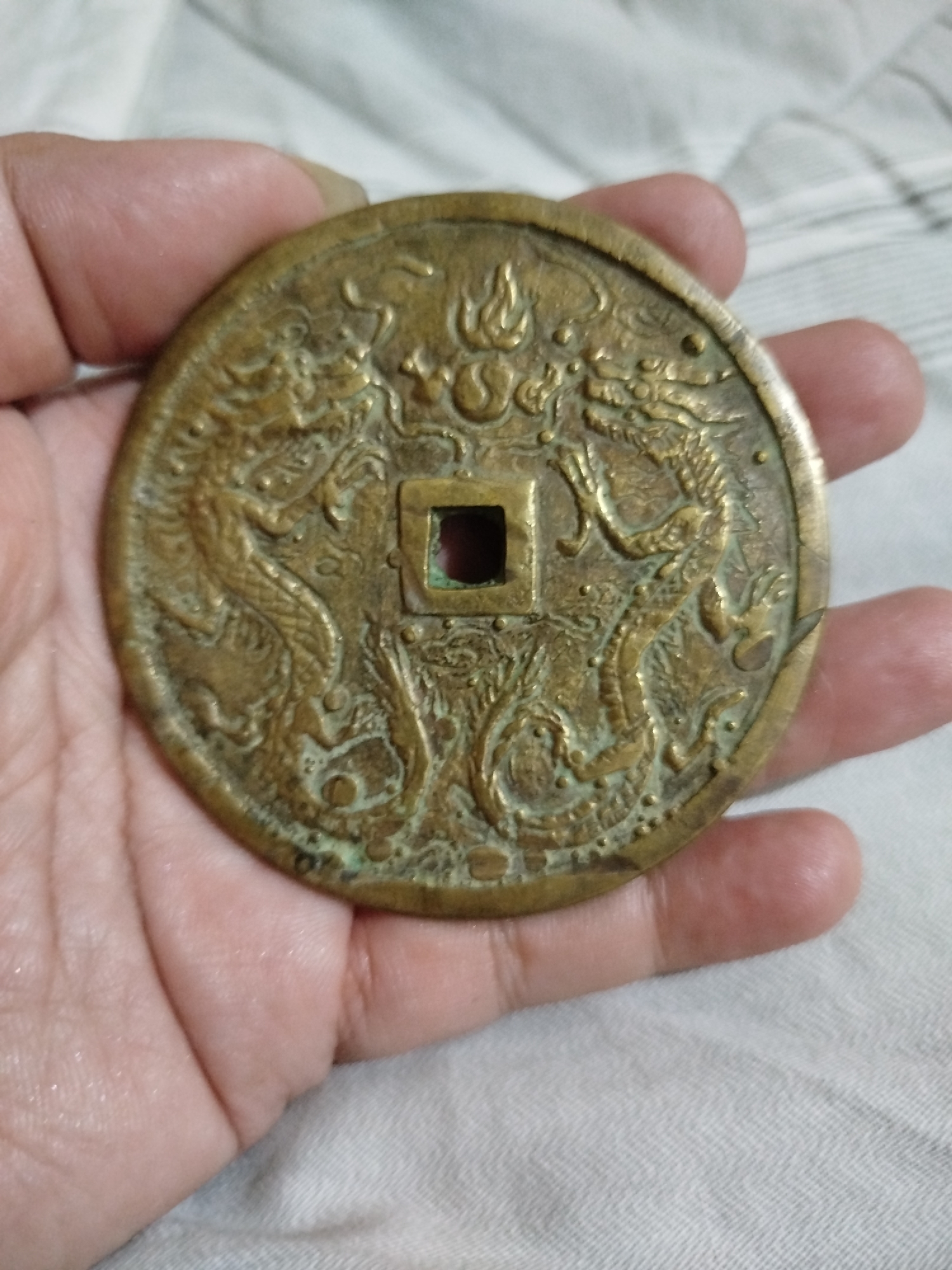 A hand holds a round, brass-colored coin with a square hole in the center. The coin is decorated with two dragons facing each other, and a flame design above the square hole. The surface of the coin appears worn and has some green patina. The background is a light-colored, textured fabric, possibly a bedsheet or blanket. There is no indication of the city of Bandungrejo, Indonesia.