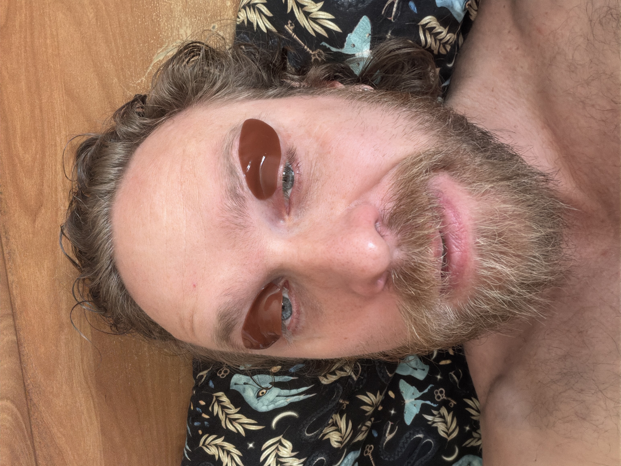 A man is lying down, with a brown cosmetic eye mask covering each eye. The man has a beard, and his hair is curly. He is lying on a patterned pillow. He appears to be on a wooden floor. The location in Saint Petersburg, Russia, cannot be determined from the image.