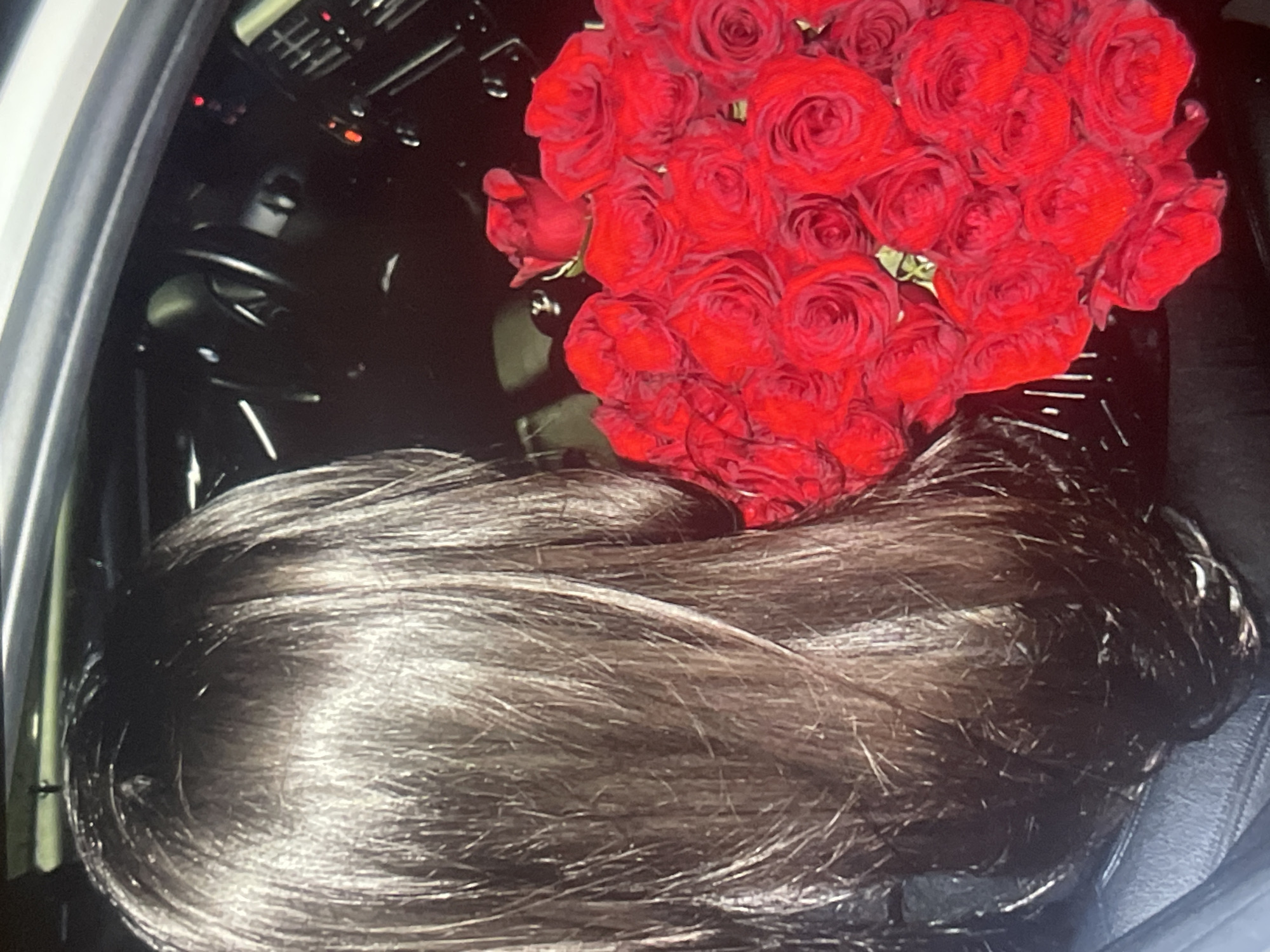 The image displays a close-up view inside a vehicle, likely a car, featuring the back of a person's head and a large bouquet of red roses. The lower half of the frame is dominated by long, dark, and shiny hair, suggesting a rich brown color with highlights from the car's interior lighting. Above the person's head, a voluminous cluster of vibrant red roses takes prominence, appearing fresh and tightly arranged.
The background reveals dim details of the car's interior, including what seems to be a dashboard or console area with air vents and faint red indicator lights. A portion of the car's door frame is visible on the far left. The overall lighting is subdued, hinting at a nighttime setting or a dimly lit interior. No specific activity is clearly depicted other than the presence of the individual and the flowers within the car. There is no visible text, and emotions cannot be discerned from this perspective. The external context for this scene is provided as Poltava, Ukraine.
