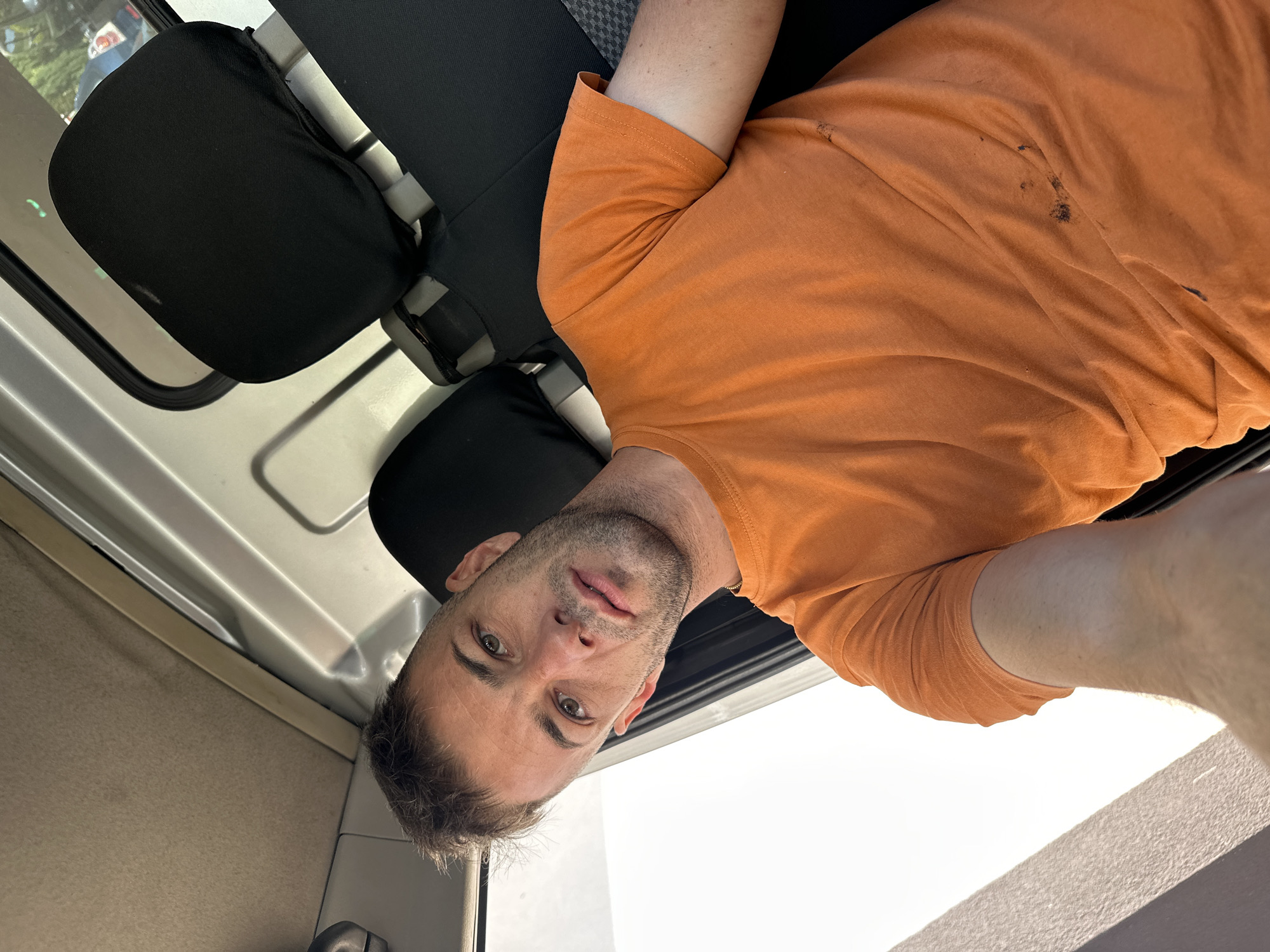 In Piperevo, Bulgaria, a man in an orange t-shirt sits in the passenger seat of a vehicle, possibly a truck, as indicated by the interior visible. The man is looking towards the camera, with his head tilted slightly. The t-shirt has dark smudges on it. The interior of the vehicle shows black seat covers and a tan headliner. The sun is bright outside the vehicle, casting a strong light on the left side.