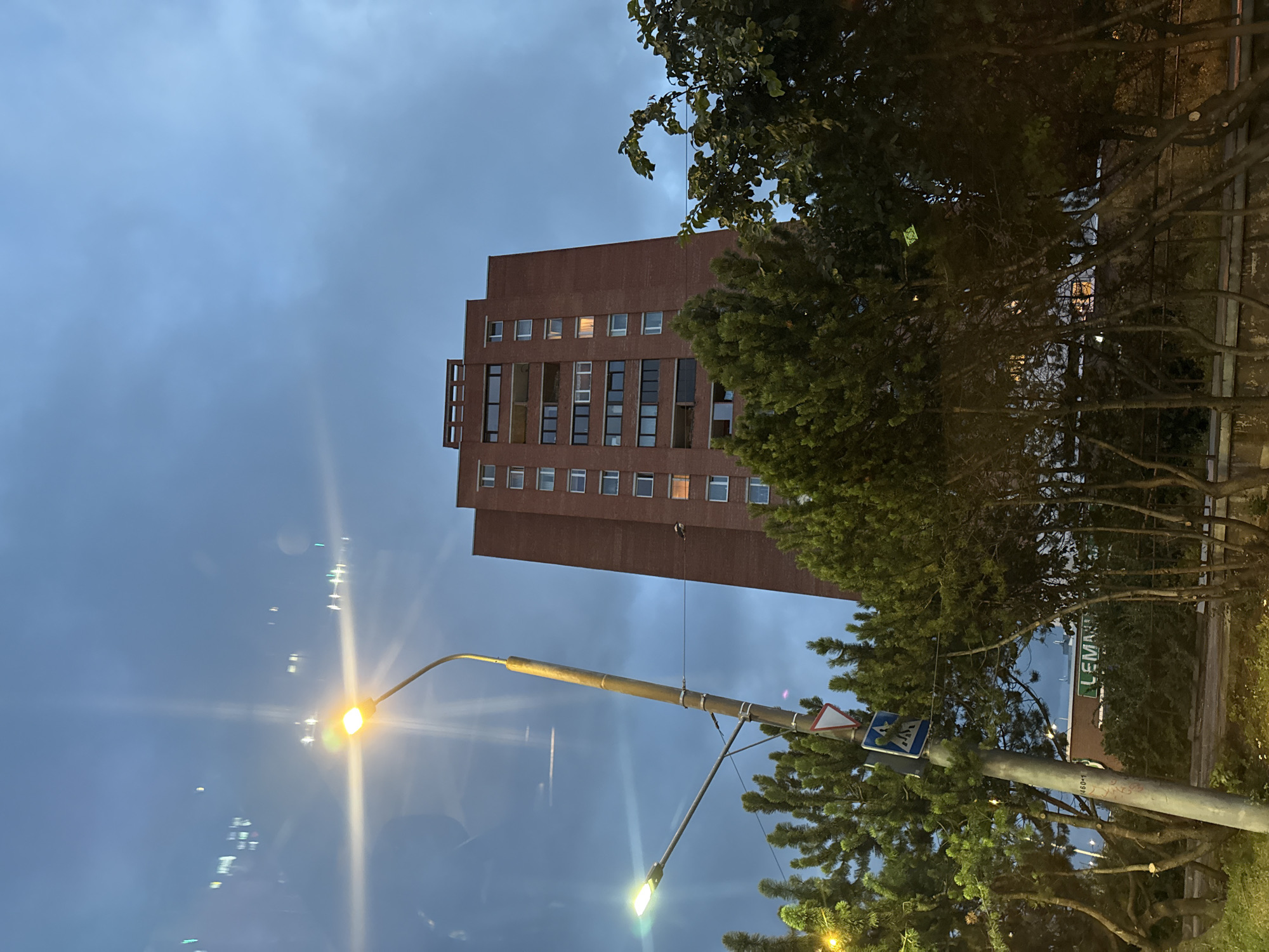 The image shows a tall, red brick apartment building in Tallinn, Estonia, under a cloudy, twilight sky. In the foreground, there are trees and streetlights, one of which is prominently illuminated. A pedestrian crossing sign is visible on a pole near the streetlight. Reflections are visible in what appears to be a window or glass surface, showing lights from other buildings. The building has many windows and balconies, and some lights are on inside. The overall scene suggests an urban environment at dusk.