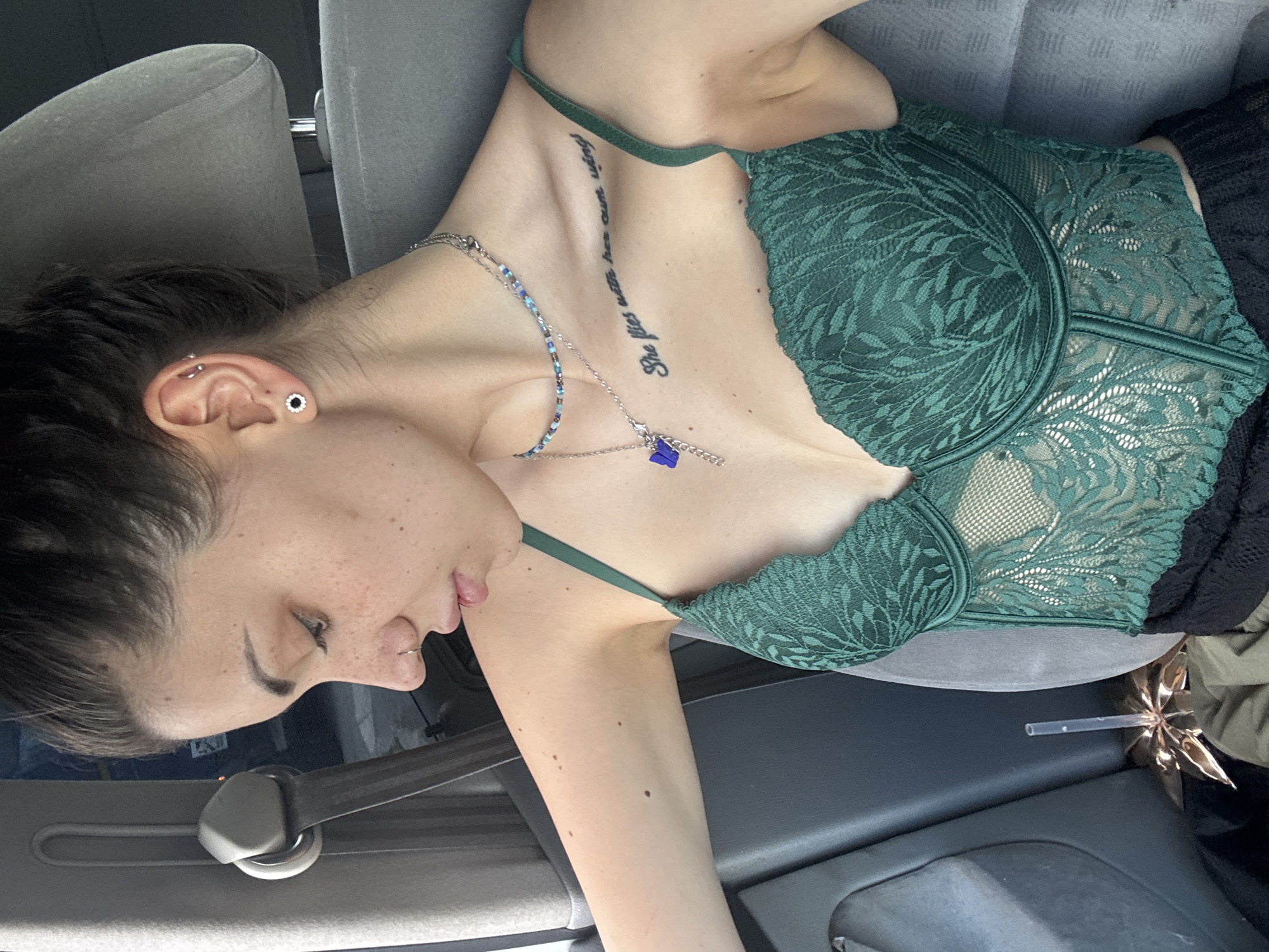 A person is shown reclining in the passenger seat of a car, likely in Elin Pelin, Bulgaria. The image is captured from an overhead perspective, with the subject lying diagonally across the frame. The person has dark hair and fair skin with visible freckles on their face and neck. They are wearing a dark green lace bralette or bustier with a distinctive leafy pattern.
Around their neck, there are two necklaces: a beaded one with blue, silver, and clear beads, and a silver chain with a small, blue, butterfly-shaped charm. On their right ear, a black and silver stud earring is visible in the lobe, and a silver hoop is in the helix. A silver hoop piercing is also visible in their nose. A tattoo on their right collarbone reads, "She flies with her own wings."
The person's eyes are mostly closed or cast downwards, and their mouth is slightly open, suggesting a state of relaxation or rest. A dark-colored garment is partially visible on the right, and a metallic gold, star-shaped object with a clear straw protrudes from between the seat and a car door, possibly a deflated balloon or decoration. The car's gray headrest and a dark seatbelt are also visible in the background, confirming the interior setting of a vehicle. The lighting suggests it is daytime.