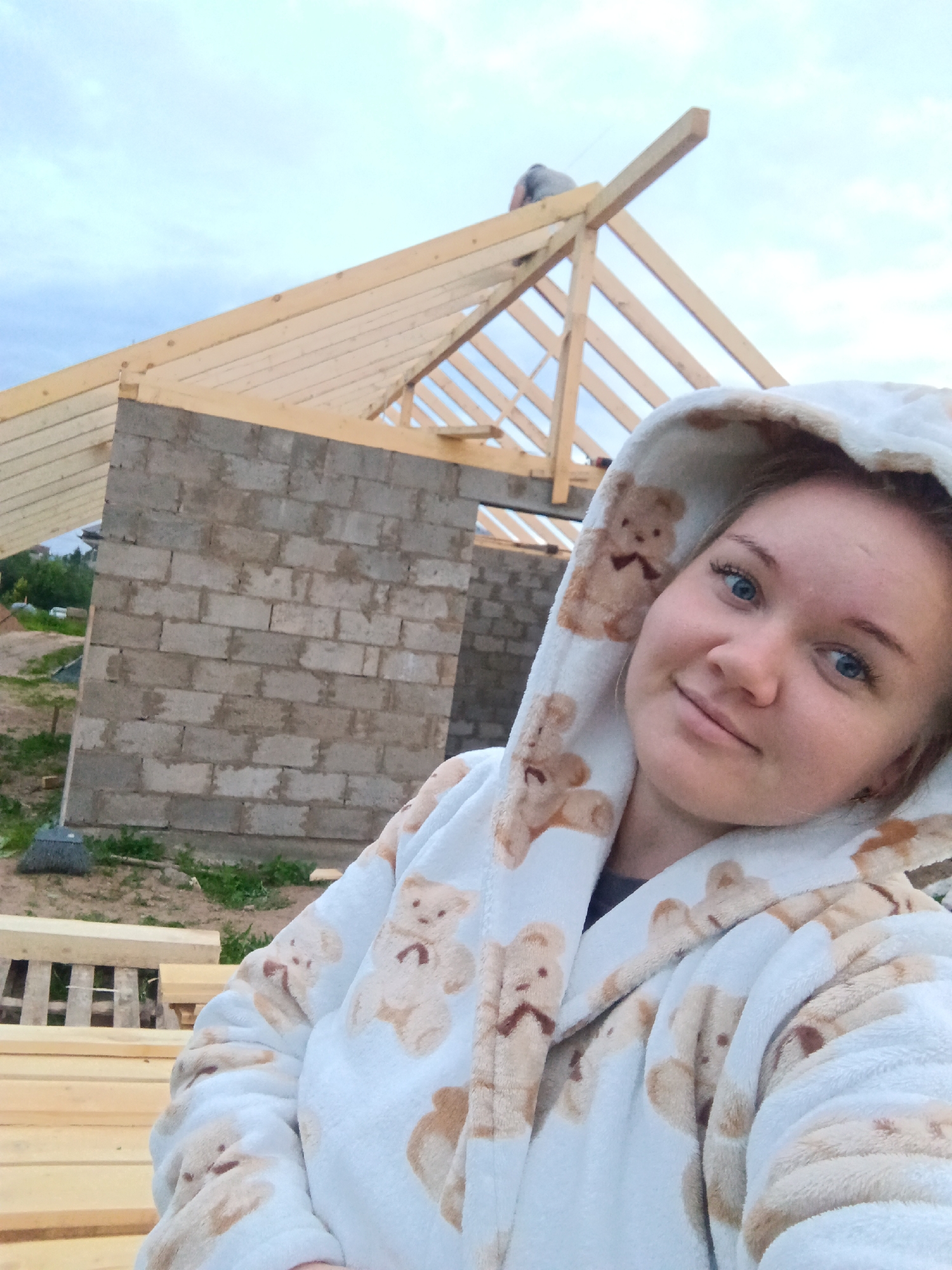 In the foreground, a young woman with blonde hair and blue eyes smiles at the camera, wearing a hooded robe patterned with teddy bears. In the background, the unfinished construction of a building is visible, with exposed wooden beams forming the roof structure and walls made of cinder blocks. The surrounding environment appears to be an outdoor construction site, with stacks of lumber in the lower left corner and a broom leaning against the wall. The sky above is overcast with a pale blue hue. The location is identified as Фоки, Russia.