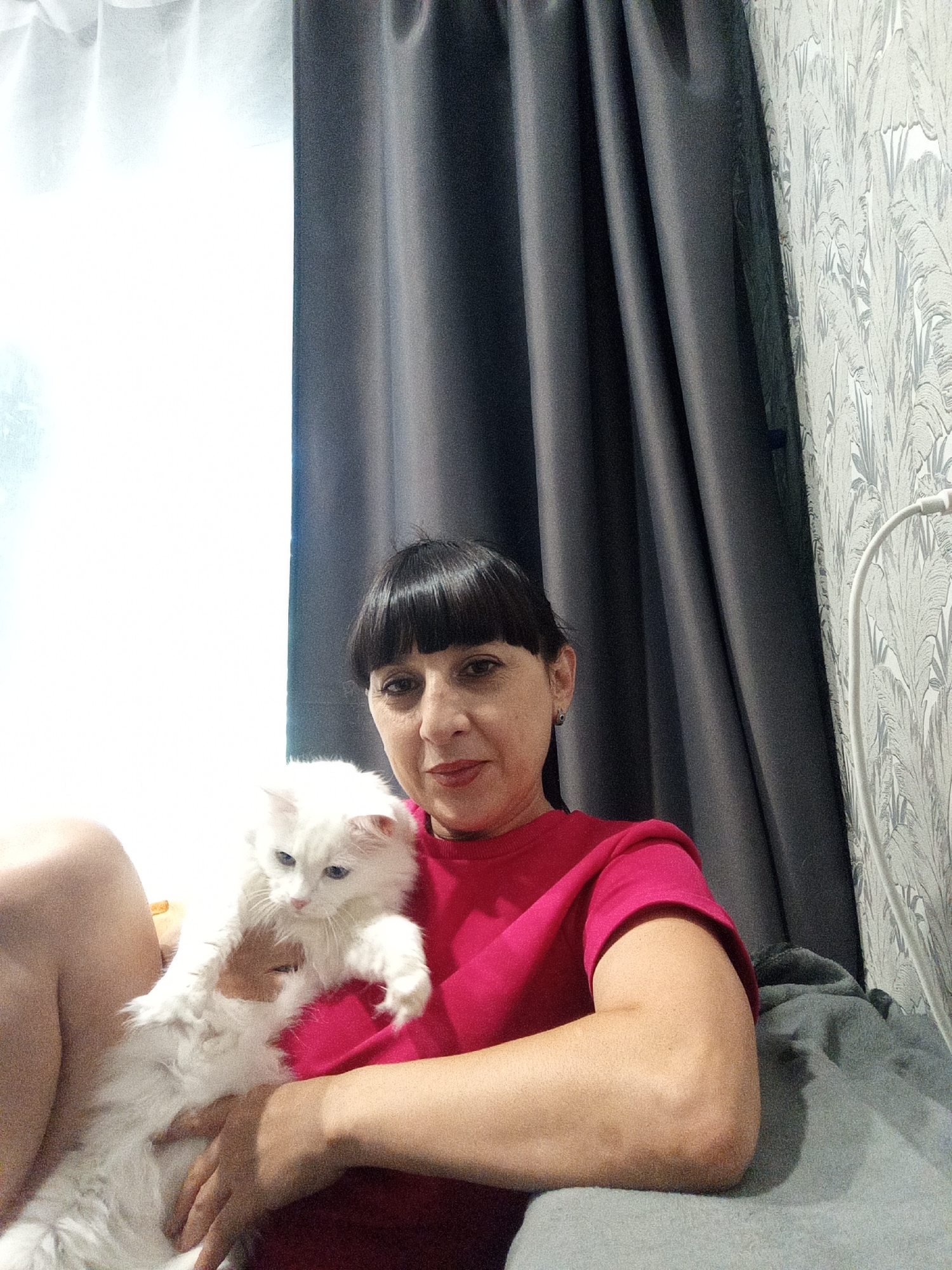 In a dimly lit room in Belogorsk, Russia, a woman with dark hair and bangs sits on a bed, holding a fluffy white cat. She is wearing a bright pink t-shirt. The cat, with blue eyes, is nestled in her arms. Behind them, dark grey curtains hang beside a window with a sheer white curtain. To the right, a wall is covered in a light-colored, patterned wallpaper, and a white lamp with a curved arm is visible. The overall impression is one of a domestic scene, possibly a home in Belogorsk.