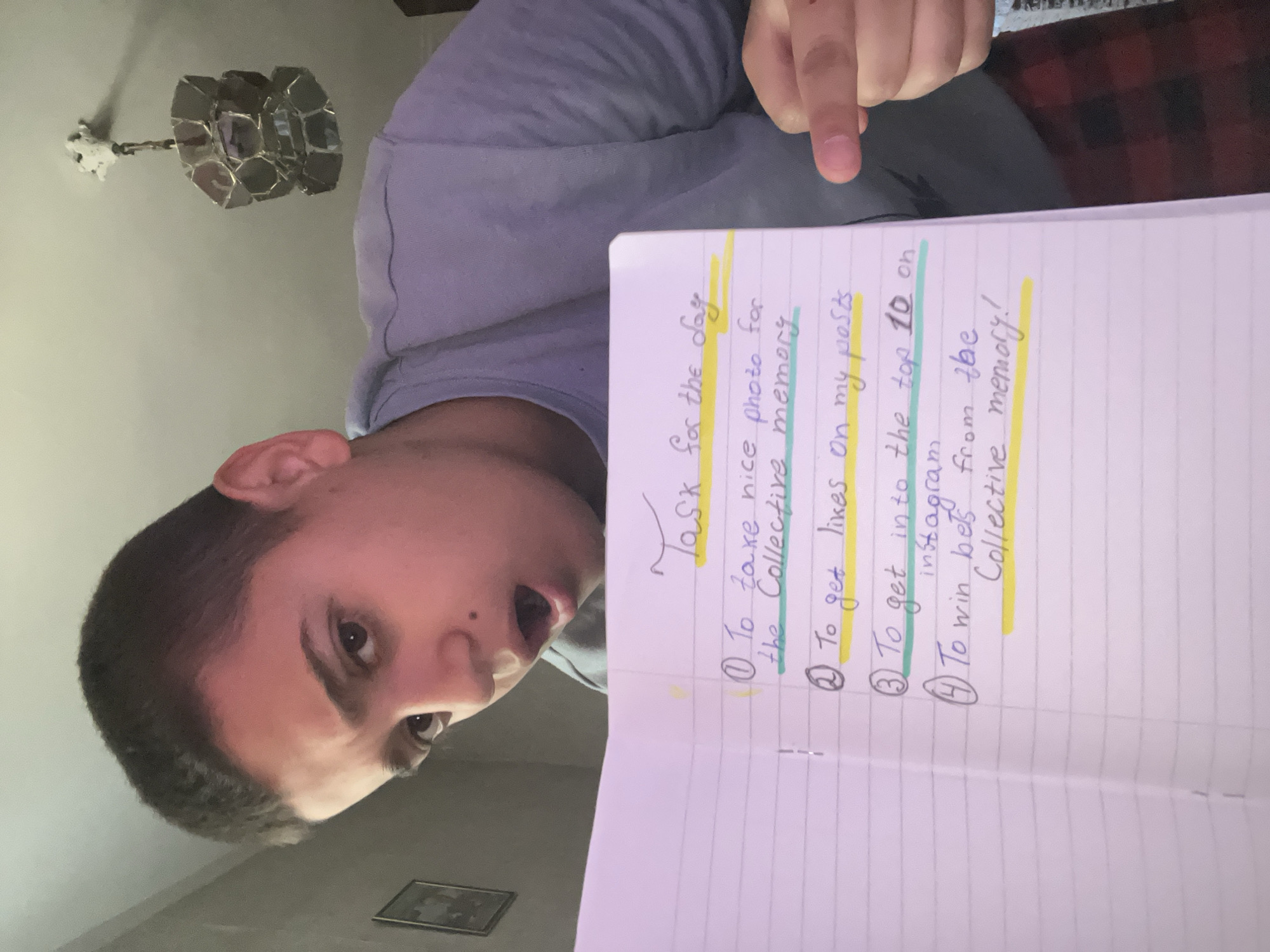 An indoor scene in Lozarevo, Bulgaria, features a young man presenting a handwritten list in a notebook. The man, with short dark hair and dark eyes, is positioned on the left, looking at the camera with his mouth slightly open, suggesting an earnest or surprised expression. His right hand, with the index finger extended, points emphatically towards the notebook from the upper right of the frame.
The central subject is an open, spiral-bound notebook filled with handwritten text, highlighted in yellow and light blue. The visible title is "Task for the day," followed by four numbered items:
1. "To take nice photo for the Collective memory"
2. "To get likes on my posts"
3. "To get into the top 10 on instagram"
4. "To win bets from the Collective memory!"
The background includes a light-colored wall and a distinctive modern light fixture on the ceiling, featuring multiple reflective, geometric components. A blurred section of furniture with a red and black plaid pattern is partially visible in the upper background, while a small framed picture adorns the wall in the lower background. The lighting is relatively dim, consistent with an indoor setting, and the overall impression is one of a personal moment focused on a set of goals or challenges.