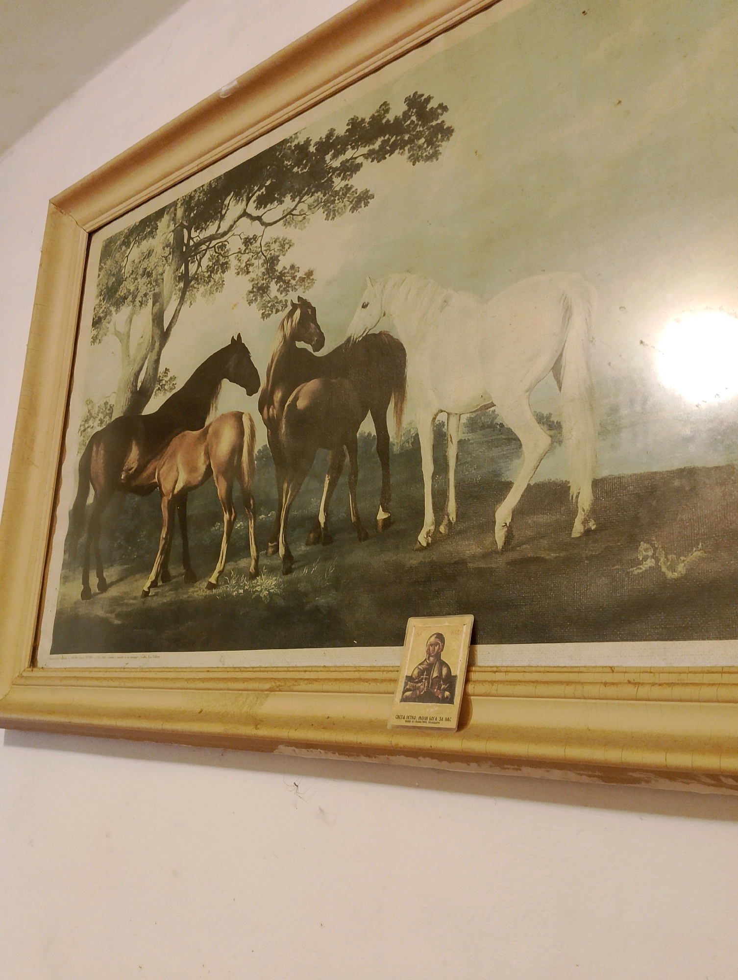 A framed painting of four horses, three brown and one white, is displayed on a white wall. The painting depicts the horses in a grassy, wooded area, with a large tree on the left side. In the foreground, a small icon of a religious figure is placed on the frame of the painting. The icon has text beneath it. The overall scene appears to be indoors, likely in a residential setting. The lighting suggests artificial light sources are present. The painting is hung at eye level.