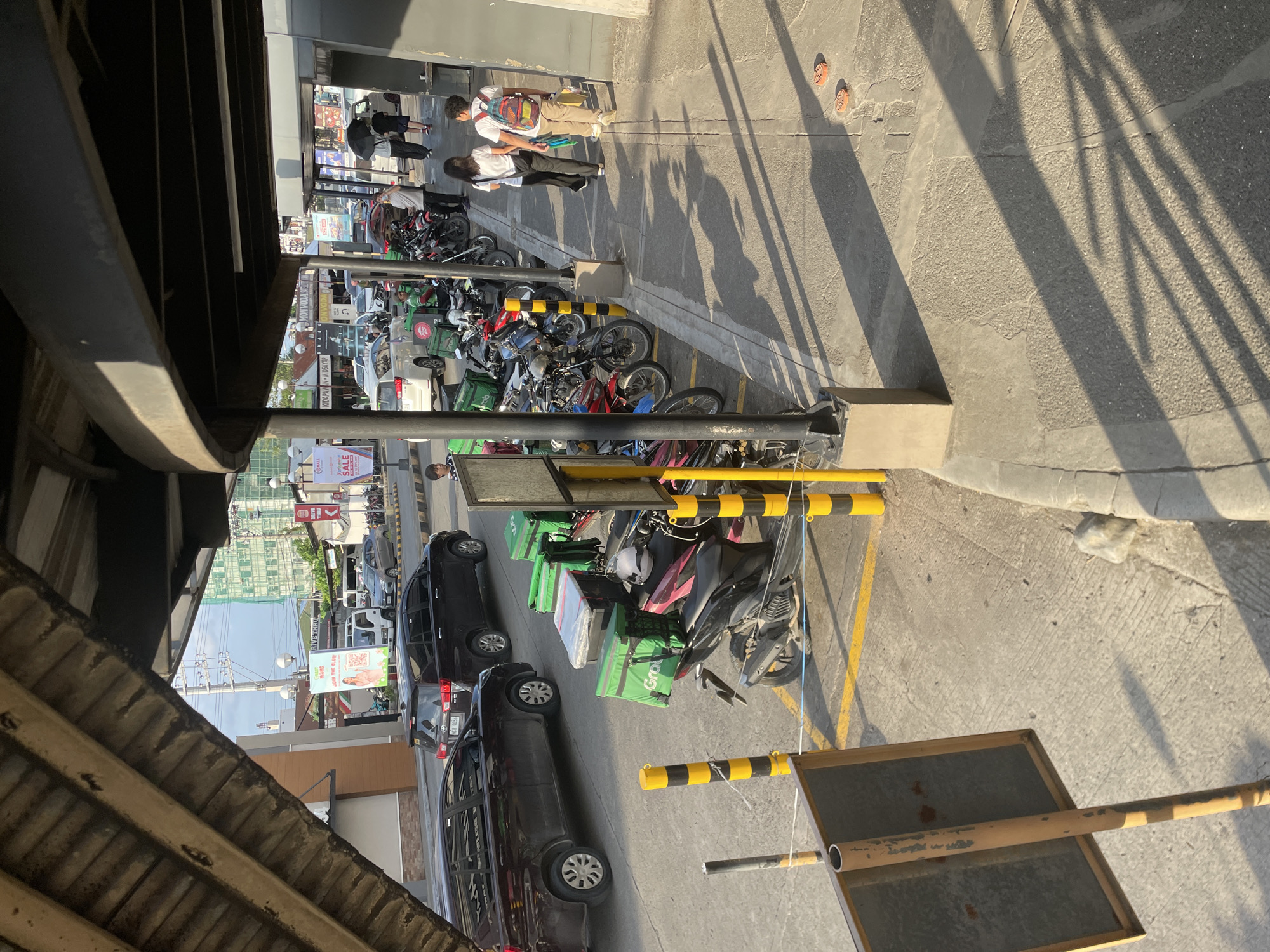 The image depicts a street scene in Davao City, Philippines, likely a commercial area given the presence of delivery vehicles and various shops in the background. In the foreground, several motorcycles with green "Grab" delivery boxes are parked along a sidewalk. Two individuals, a woman in black pants and a white top and a man with a backpack and light-colored pants, are walking on the sidewalk away from the viewer, casting long shadows. Another person, holding a black umbrella, walks ahead of them. Behind the motorcycles, there are cars, including two dark SUVs, parked on the street. In the mid-ground, a white delivery truck with "Grab" branding is visible. Signs for "SALE" and "DRIVE THRU" are present in the background, suggesting a retail or dining establishment. The overall environment is characterized by a concrete pavement, sunlight creating distinct shadows, and a mix of vehicles and pedestrians.