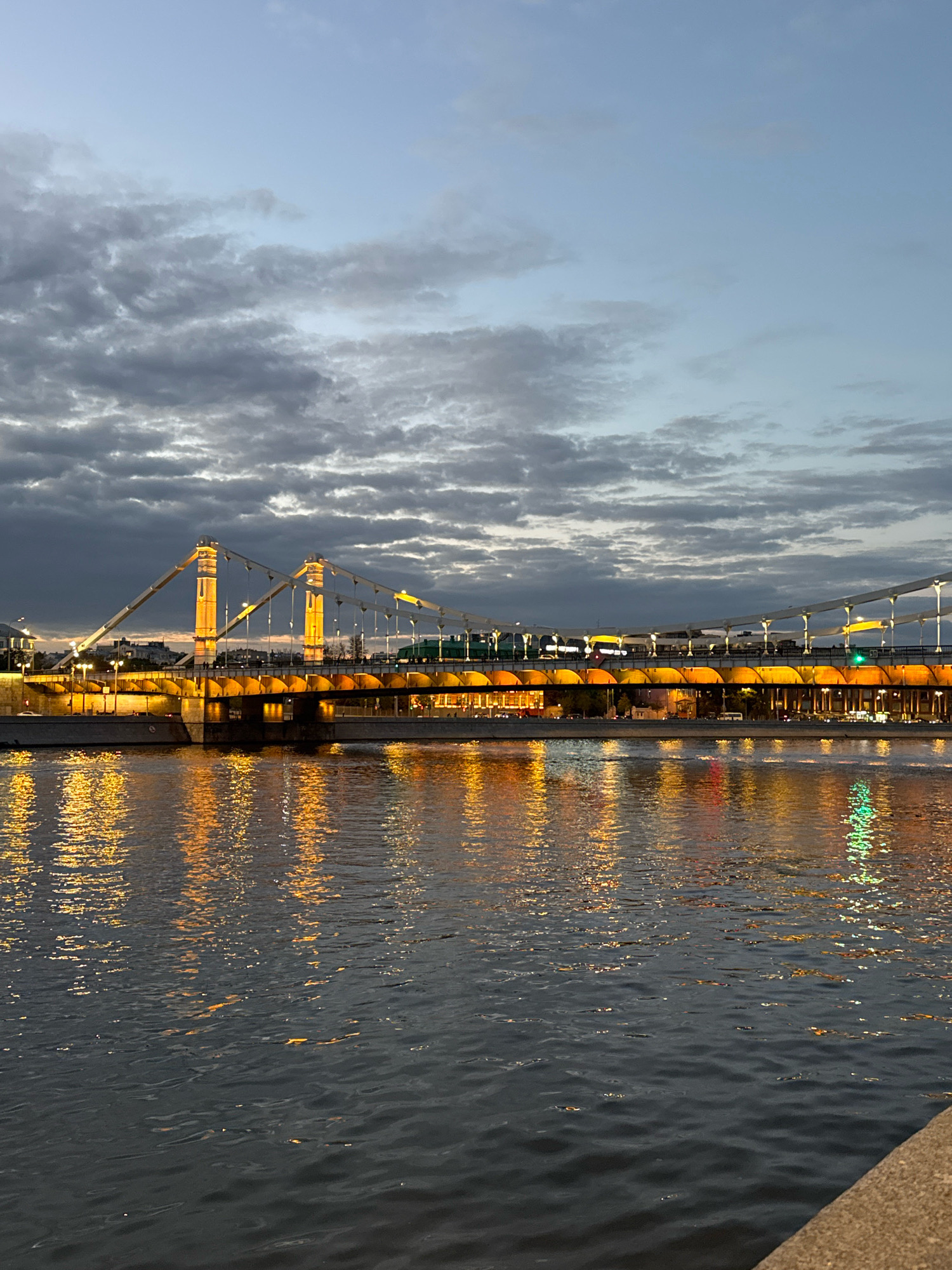 The image shows the Krymsky Bridge in Moscow, Russia, at dusk. The bridge is illuminated with golden lights, and its reflection is visible in the Moskva River. The sky is cloudy, with patches of blue showing through. There are some vehicles visible on the bridge, and the water is rippling. The overall mood of the image is serene and picturesque.