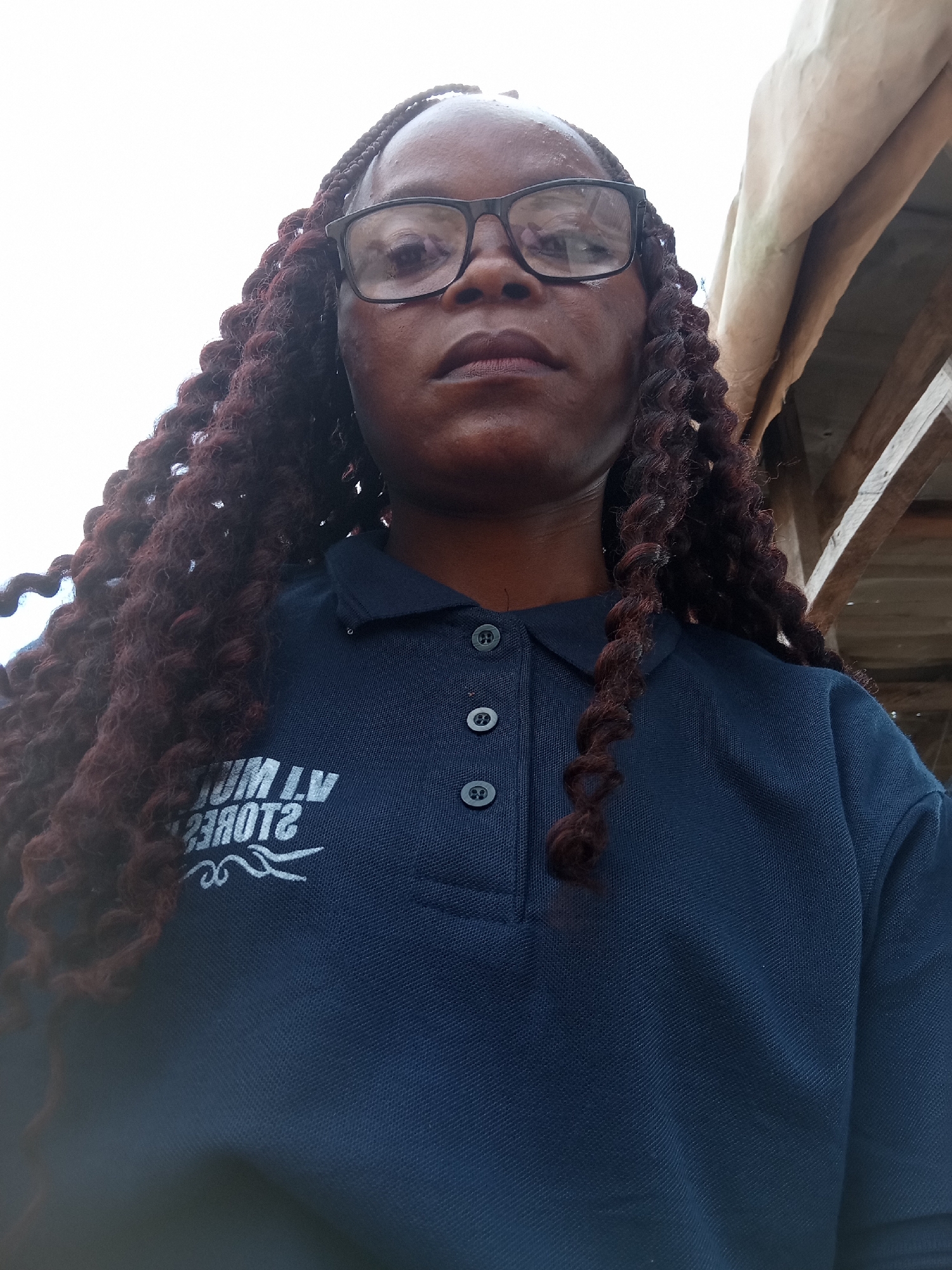 A low-angle, close-up, indoor selfie captures a Black woman from the chest up, wearing glasses and a navy blue polo shirt with a white logo on the left chest. She has dark, braided hair that extends down past her shoulders. The woman's expression is neutral. The background consists of the white sky and a beige tarp supported by wooden beams, suggesting an outdoor or semi-outdoor setting under a shelter. The logo on her shirt appears to be a reversed text that reads "V.M. MULTI STORES" along with a stylized design. The overall scene is ordinary, with no discernable location markers pointing to Jalingo, Nigeria.