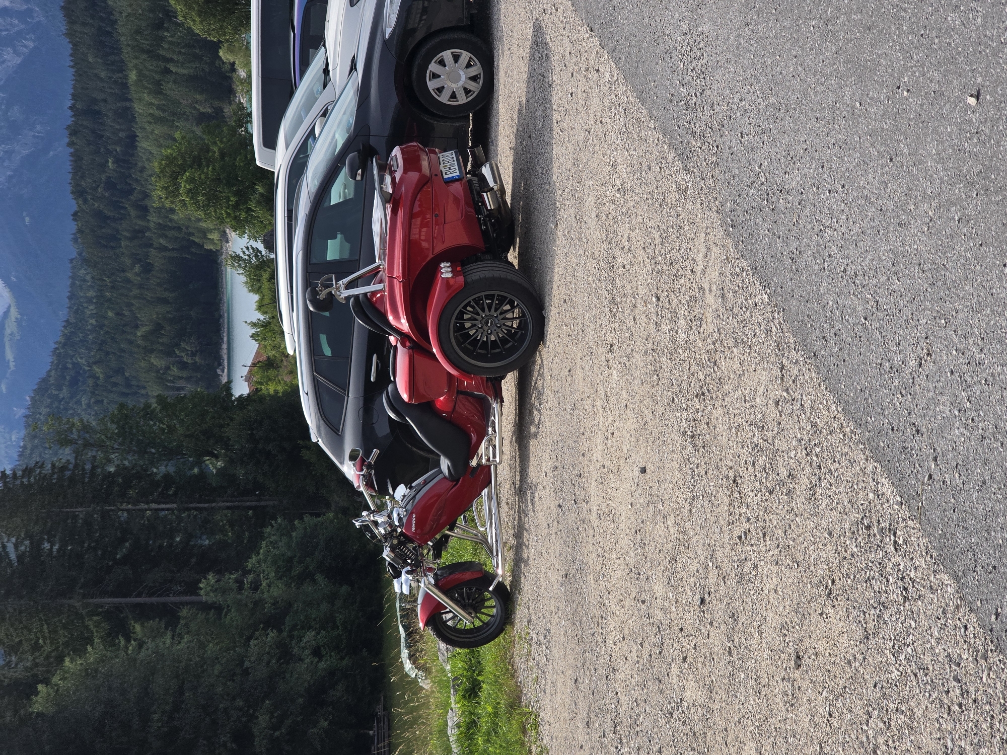 In Grainau, Germany, a red three-wheeled motor vehicle, resembling a trike, is parked on a gravel lot next to several cars. The trike has a chrome frame, a black leather seat, and a license plate that reads "RHB002". Behind the trike and cars, a dense forest of pine trees covers a hillside, leading up to a body of water and distant mountains. The scene suggests a scenic outdoor location, possibly a parking area for tourists visiting the natural landscape. The lighting indicates it is daytime, with the sun casting shadows. No people are visible in the frame.