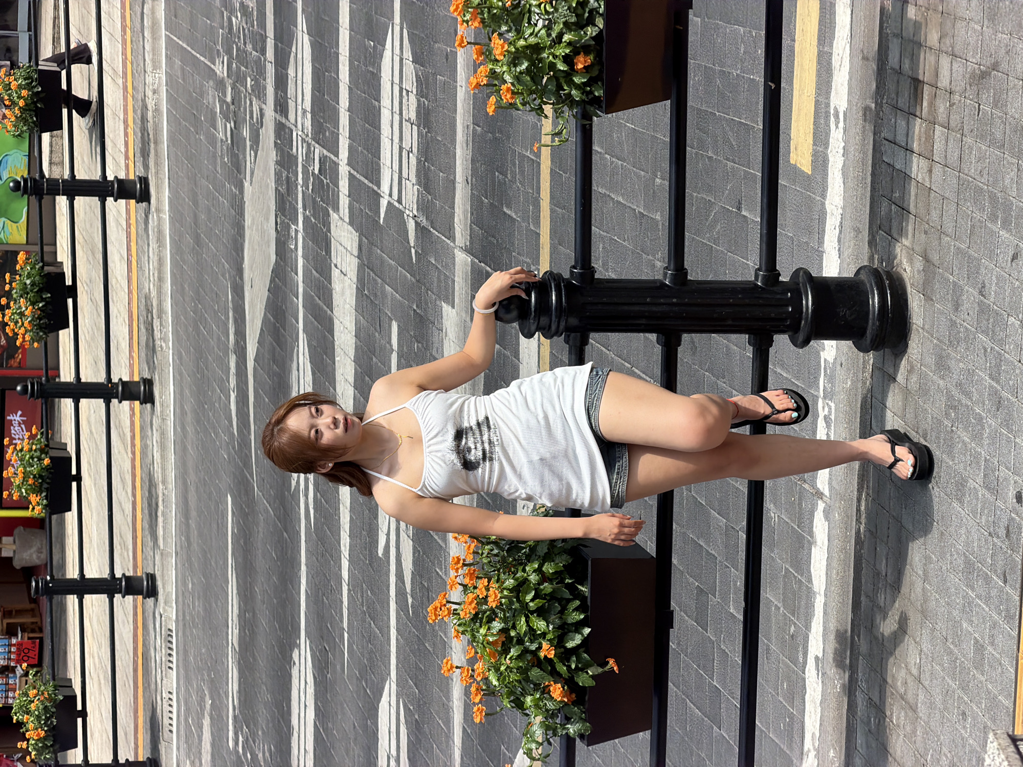 A young Asian woman stands on a tiled pedestrian walkway in Yuzhong District, China, leaning against a black metal railing. She is wearing a white halter-neck tank top with a graphic print and denim shorts. Her hair is light brown and pulled back into a ponytail. She has on black platform flip-flops with light blue toenail polish. In front of her, on the walkway, are two black rectangular planters filled with vibrant orange flowers. Behind the railing, the ground appears to be a road with white and gray striped crosswalk markings. A person in dark pants and white shoes is visible walking in the background. The lighting suggests it is daytime with strong sunlight, casting distinct shadows.