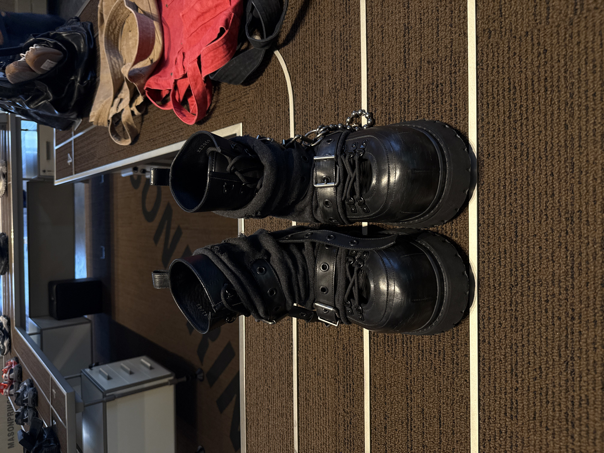 The image shows a pair of black leather boots with buckles and chains on a brown textured carpet. The boots are positioned parallel to each other and are placed near a white line. In the background, there are pieces of clothing, including a red garment and a tan belt, placed on what appears to be a shelf. In the far background, there is what appears to be a retail display with various items, including other shoes. There is no indication of the location being the Liwan District in China.