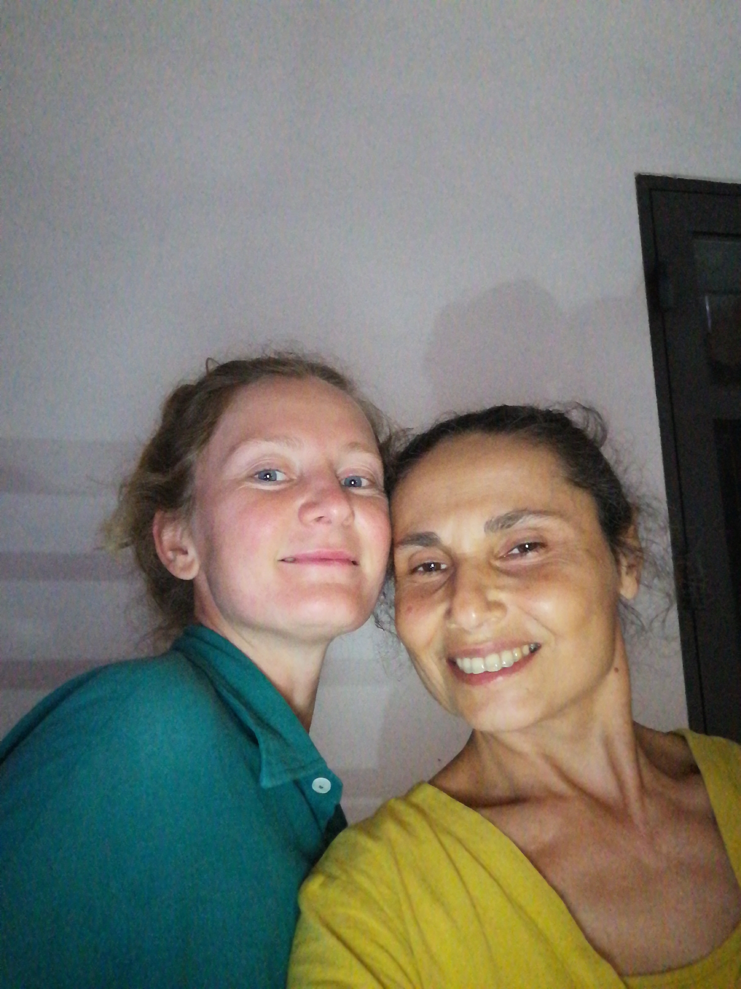 The image is a selfie of two women. The woman on the left has fair skin, blue eyes, and her blonde hair is pulled back. She wears a teal collared shirt. The woman on the right has darker skin and dark hair pulled back. She is wearing a yellow top and smiling, showing her teeth. They are close together, possibly touching.
The background is a plain white wall, and there is a dark object in the upper right corner, possibly a doorway or window frame.
Given that you have specified the location, it is implicitly understood that this photograph was taken in Bai Chay Ward, Vietnam, though there are no visual cues that confirm this in the image itself.