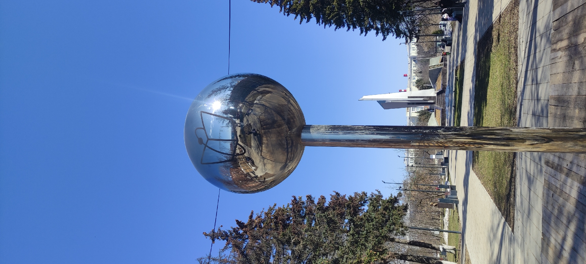 The image shows a reflective spherical sculpture mounted on a pole in a park-like setting under a clear blue sky. The sculpture reflects the surrounding environment, including trees, buildings, and the sun. A thin wire is visible near the top of the sphere.
The location is Fryazino, Russia, based on the sculpture and surrounding architecture. A sidewalk runs alongside a grassy area with benches. Several people can be seen in the reflection within the sphere. The base of the sculpture sits on a wooden platform. The sun is reflecting off of the sphere. A modern building can be seen in the background.