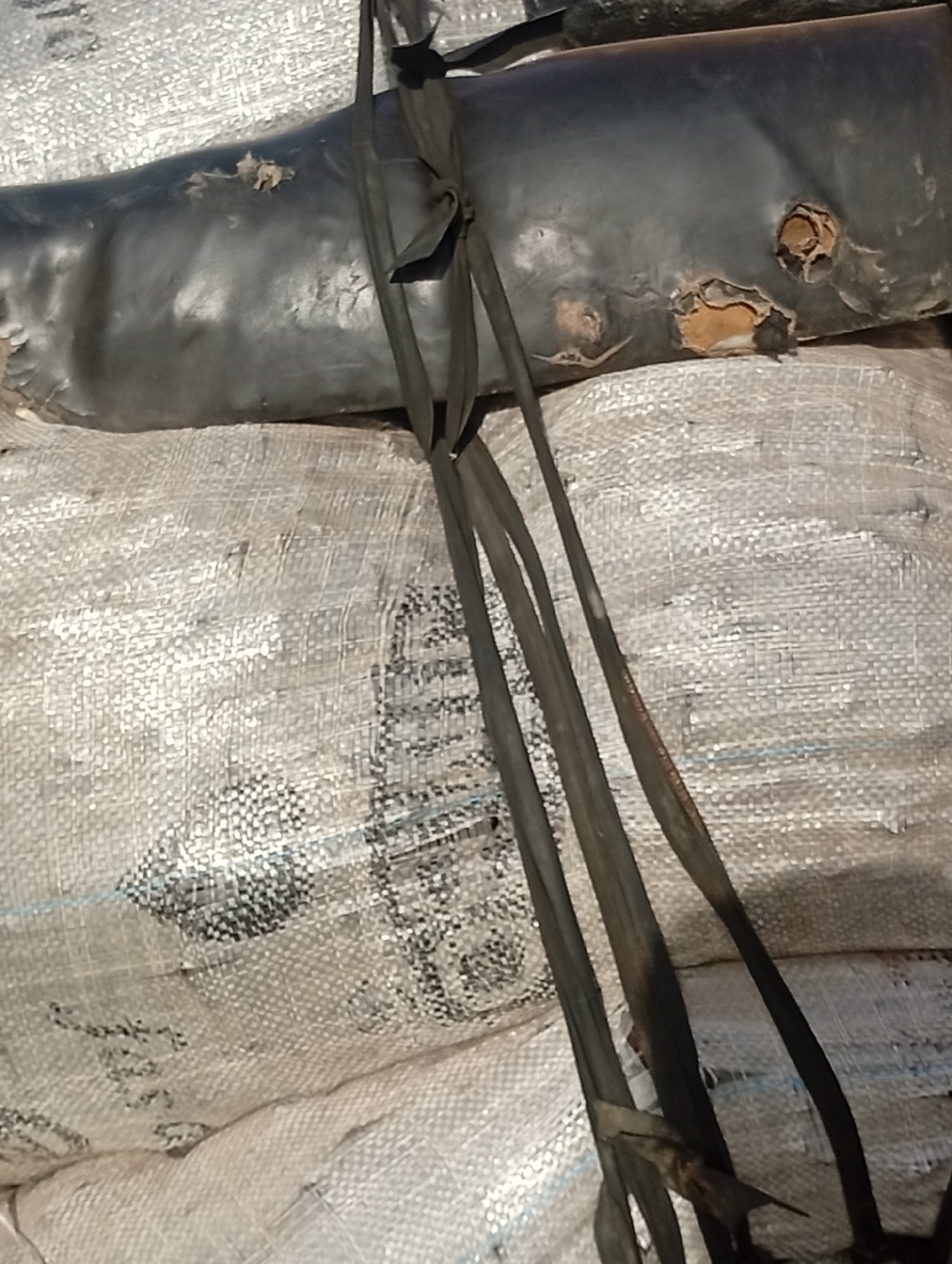 The media file shows a close-up view of what appears to be a damaged, black, synthetic material object resting on top of large, woven sacks. The black object is dark and appears to be somewhat deflated or compressed, with several visible tears and abrasions that reveal a lighter, possibly tan-colored material underneath. Black straps or bands are attached to the black object and extend downwards, some of which are tied in knots.
The woven sacks beneath the black object are light-colored and have a textured surface. Some portions of the sacks show faint dark patterns or text, though it is not clearly legible. The overall impression is one of discarded or damaged materials, possibly in an outdoor or storage setting. The lighting suggests it is daytime, but there are no specific cues about the time of day or weather conditions. The image is a still photograph and does not depict any activity or event taking place. The focus is entirely on the texture and condition of the materials.