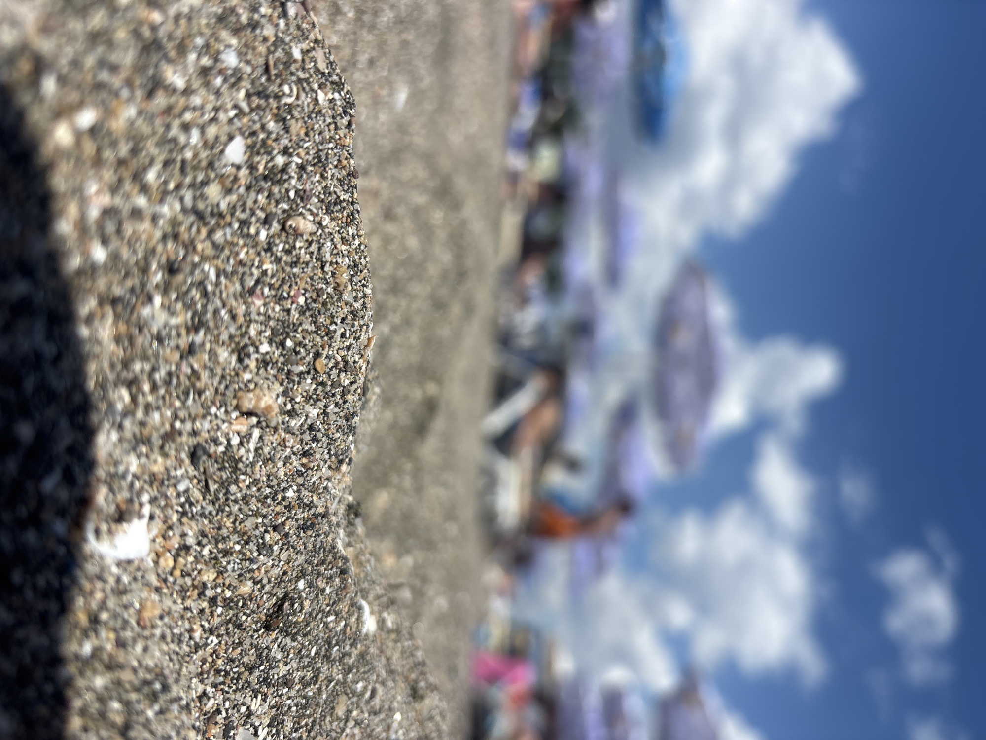 A low-angle, close-up shot of wet sand on a beach shows small pebbles and shells. In the background, out of focus, are beach umbrellas, lounge chairs, and people enjoying a sunny day under a blue sky with white clouds.