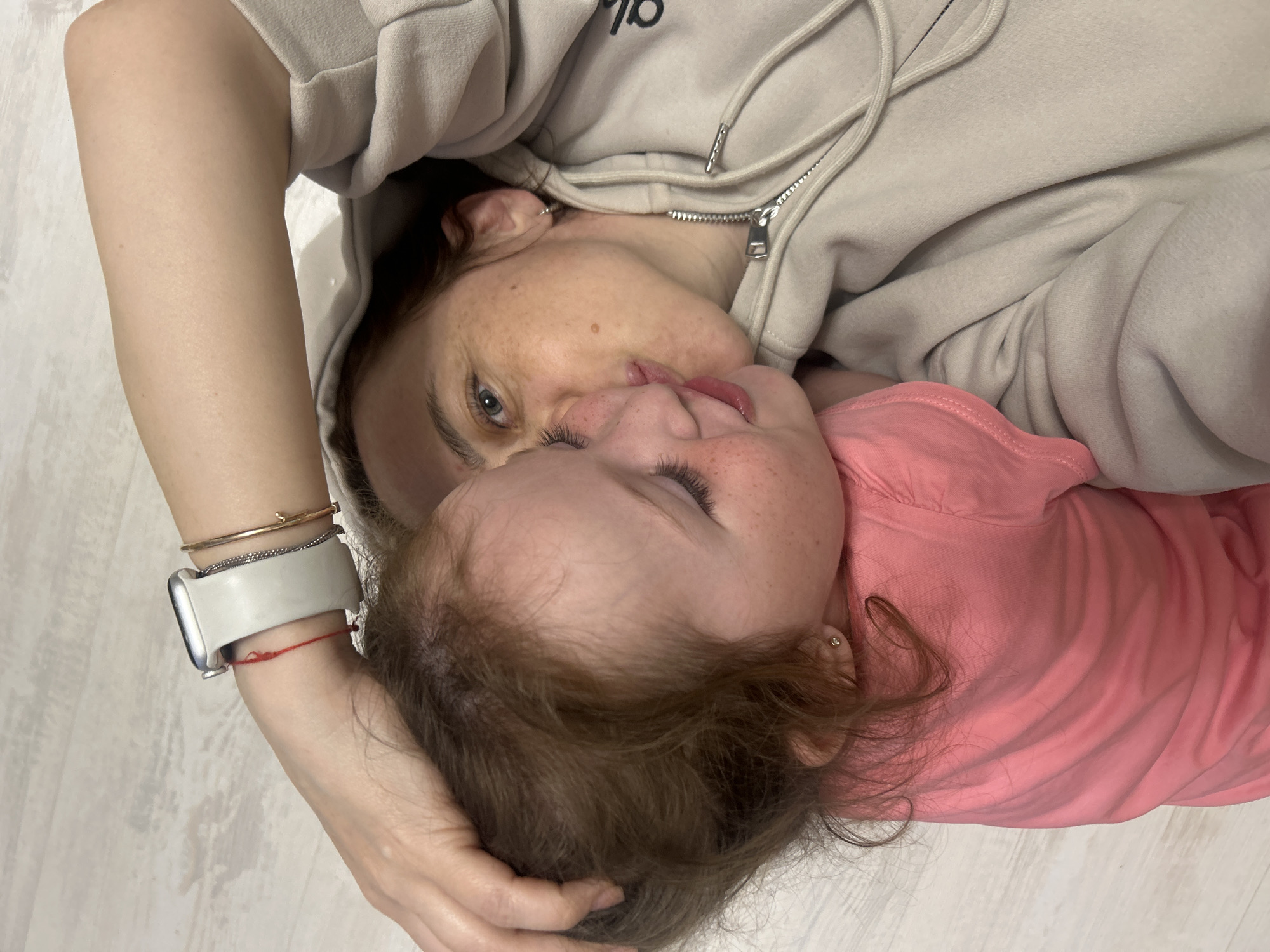 The image shows an adult and a child lying down. The adult is wearing a light-colored hoodie with a zipper. The child is wearing a pink shirt. Their faces are close together, and their tongues are touching. The adult is wearing a watch and bracelets on their wrist. The background appears to be a light-colored floor. The image does not provide enough information to determine if this photo was taken in Chaikovsky, Russia.