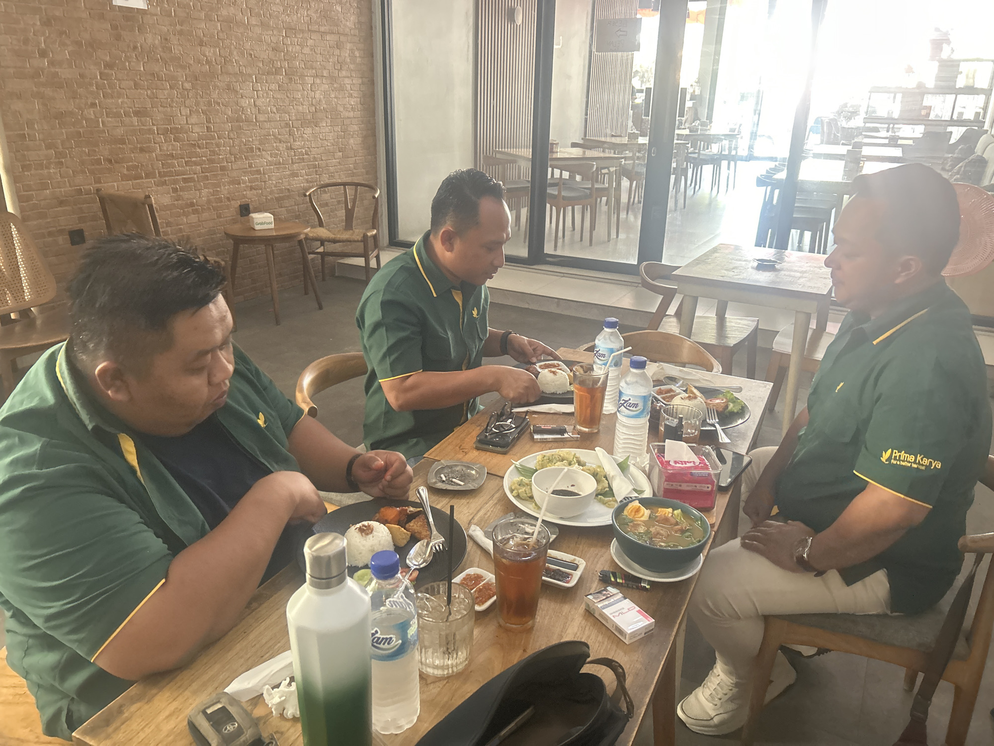 Three men in green shirts with yellow trim are seated at a wooden table in a cafe, likely in Brebes, Indonesia, based on the "Prima Karya" logo on their shirts, which is a known company in that region. The man on the left, with a dark shirt underneath his green one, is eating from a plate of rice and fried items. The man in the center is also eating. The man on the right is facing away from the camera, looking at his food. On the table are plates of food, including rice dishes and a bowl of soup, several bottles of water, glasses of iced tea, cigarettes, an ashtray, and other condiments. The cafe appears to have a rustic interior with exposed brick walls and wooden furniture. Large windows in the background reveal more tables and chairs, suggesting a spacious dining area. The overall atmosphere is casual and relaxed, with the men engaged in a meal.