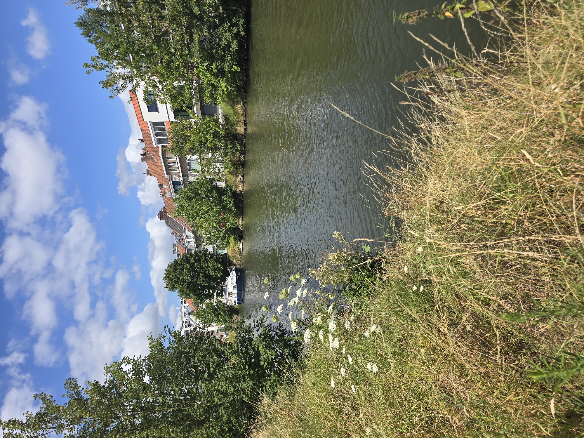 This is a photograph of a canal in Ghent, Belgium. The canal is flanked by lush green trees and a grassy bank in the foreground. Across the canal, a row of buildings with terracotta tiled roofs and modern white facades are visible, some with balconies. A white boat with a dark blue stripe is docked on the canal, and a smaller white object, possibly another boat or dock, is visible further down the waterway. The sky is a vibrant blue with scattered white cumulus clouds. The scene is peaceful and appears to be taken on a sunny day.