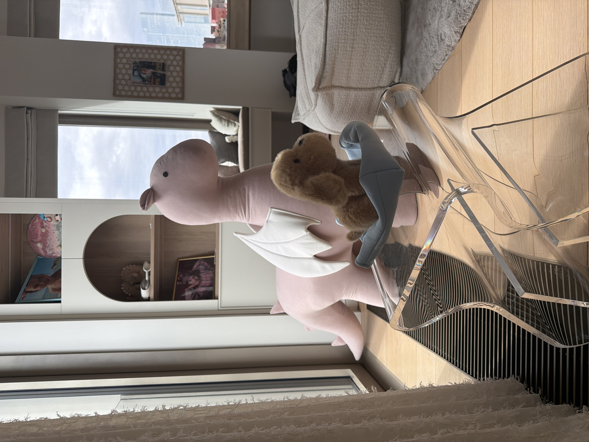A room interior is visible, featuring a pink, winged stuffed dragon with a stuffed bear perched atop it. The dragon and bear sit on a transparent acrylic chair with a slatted base, positioned near a window. Outside the window, a cityscape with skyscrapers is partially visible. The room also contains a white couch, a light-colored wooden floor, and built-in shelving with various items displayed. It is not possible to determine the location as Moscow, Russia, from the given information.