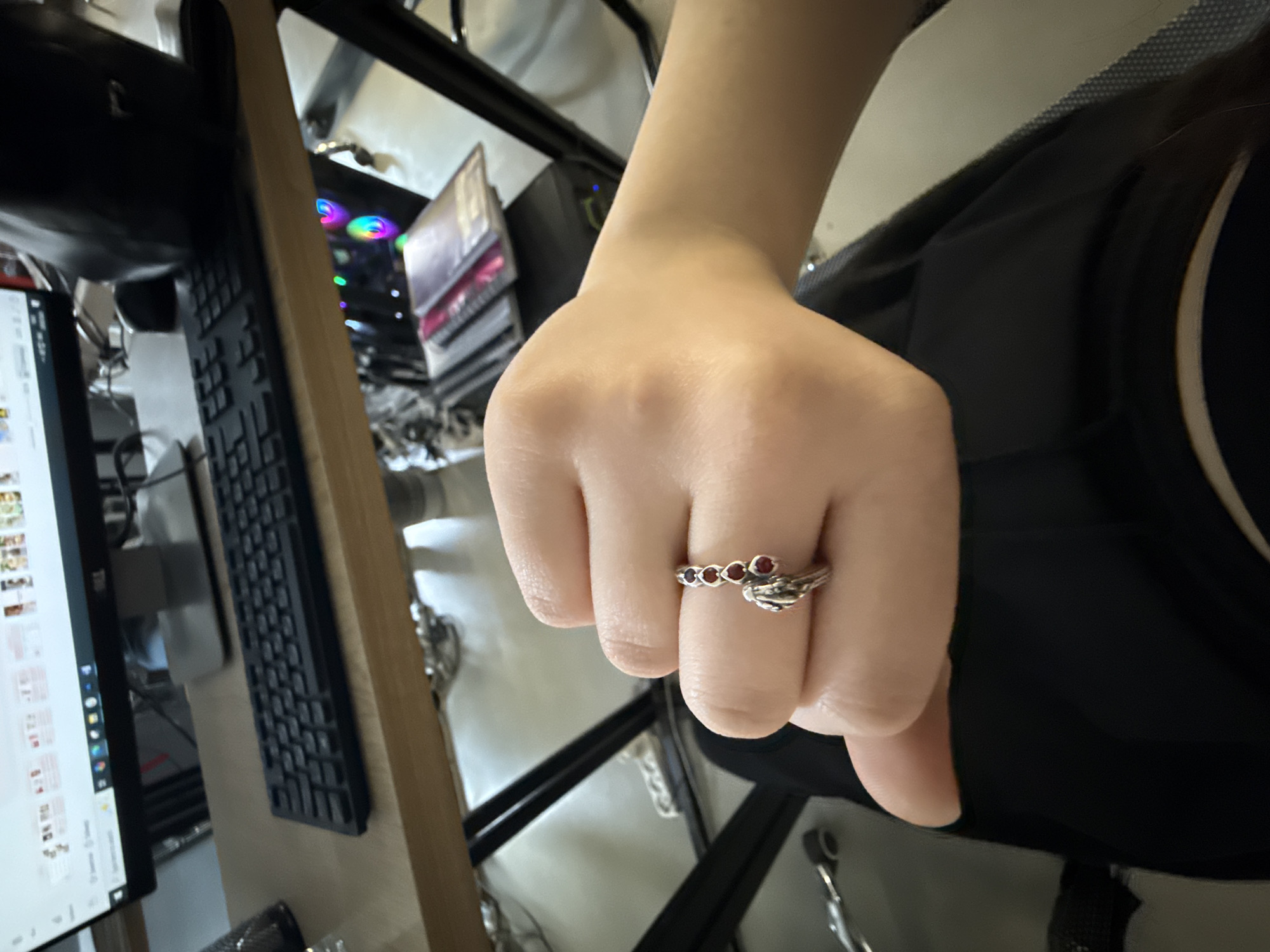 A fair-skinned human left hand, with fingers slightly curled, is prominently centered in the foreground. A silver-toned ring is worn on the third finger, featuring a linear arrangement of five small, red, possibly heart-shaped stones, and a stylized, organic silver element, resembling a leaf or feather, extending towards the palm. The subject is wearing dark, likely black, clothing visible beneath the hand.
The background shows an indoor workspace. A light brown wooden desk surface occupies the upper half of the image. On the desk, a black computer monitor, branded "DELL," displays a graphical user interface with multiple tabs containing various small image thumbnails. A black computer keyboard is positioned in front of the monitor, and a black computer mouse and a black speaker or headphone stand are also present. Underneath the desk, a computer tower is visible, emitting colorful RGB lighting in hues of blue, green, and pink/purple from its internal components. Several binders or notebooks are stacked near the computer tower. The desk framework is constructed of black metal tubing. The immediate surroundings lack specific identifying markers for the city of Yerevan, Armenia.