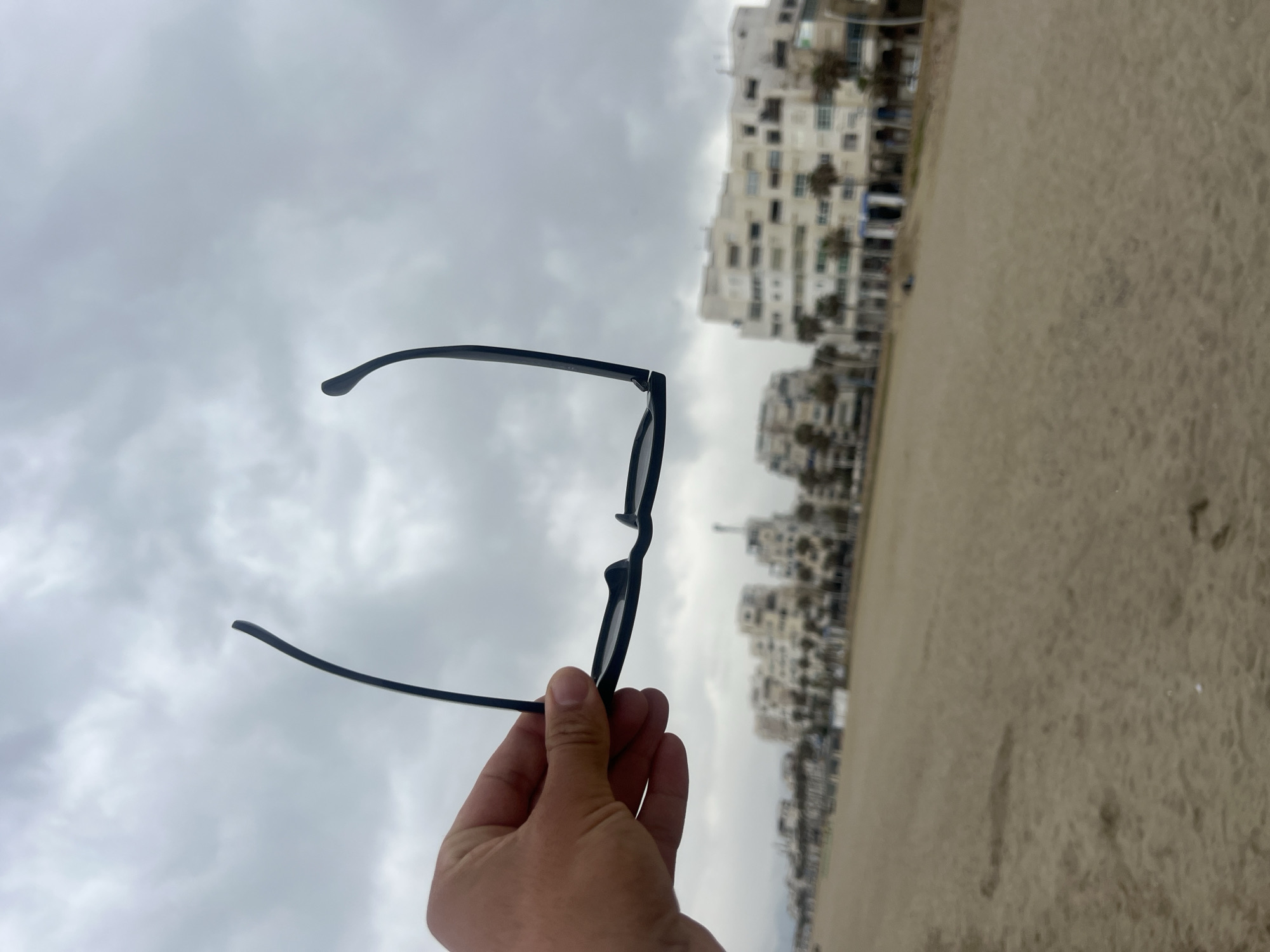 The image, when viewed in its correct orientation, depicts a person's hand holding black-framed glasses against a coastal backdrop on an overcast day.
In the foreground, a light-skinned hand, presumably a right hand, holds a pair of dark-colored, possibly black, framed glasses. The glasses are held by one of the temple arms and part of the frame, with the arms partially folded. On the inside of the visible temple arm, faint white text "CE" is discernible.
The background features a wide, light-brown sandy beach occupying the lower portion of the scene. Stretching along the horizon, beyond the sand, is a row of multi-story buildings. These structures are predominantly light-colored, possibly white or beige, with many windows, suggesting apartment complexes or hotels typical of a coastal urban area. The sky above is entirely covered with thick, gray clouds, indicating an overcast or cloudy day with soft, diffuse lighting. There is no direct sunlight visible. The setting appears to be a beach in a developed coastal town, consistent with Martil, Morocco. The time of day is daytime, but the weather is notably cloudy.