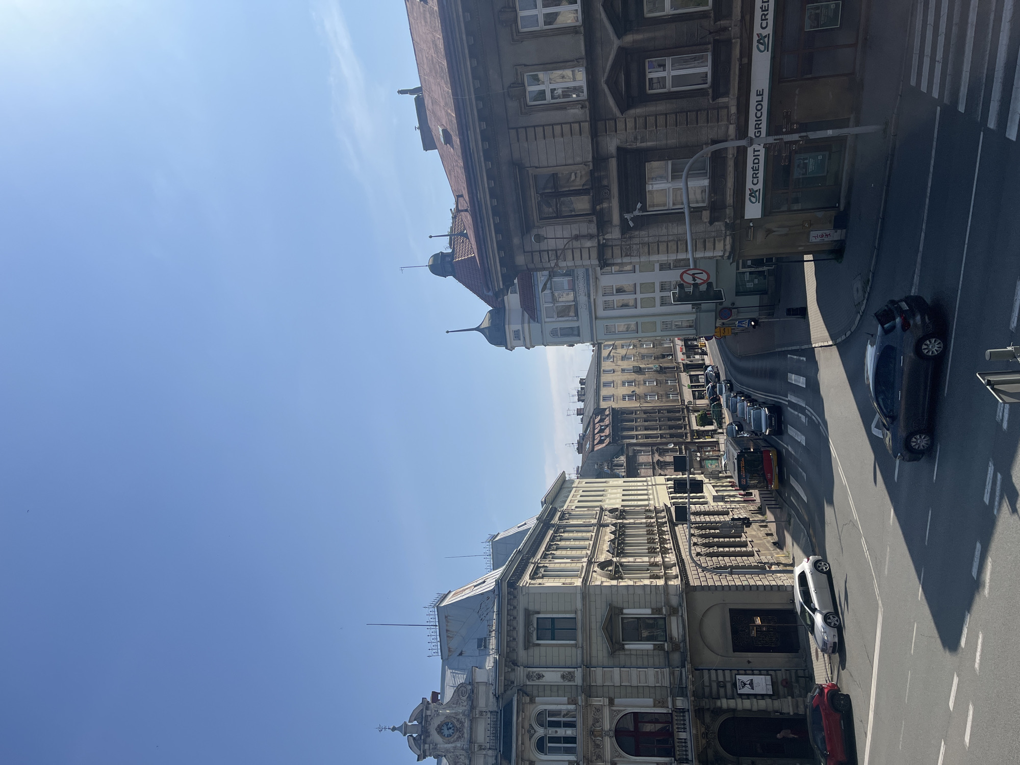 The image shows a street scene in a European city with vintage architecture. The sky is clear and blue. On the left, a light-colored building with ornate carvings and a clock tower is visible. Adjacent to it is a building with a mansard roof and arched windows. On the right, a darker, older building with a rough facade and multiple windows stands. Further down the street, other buildings of varying heights and architectural styles line the road. Several cars are present on the asphalt road, including a red SUV, a silver sedan, and a dark-colored hatchback. A yellow and red bus with "22" displayed on its front is also stopped at a traffic light. A pedestrian is visible near the entrance of a building on the left. Traffic lights and street signs are also present. A crosswalk is marked on the road. The overall scene depicts a moderately busy urban environment.