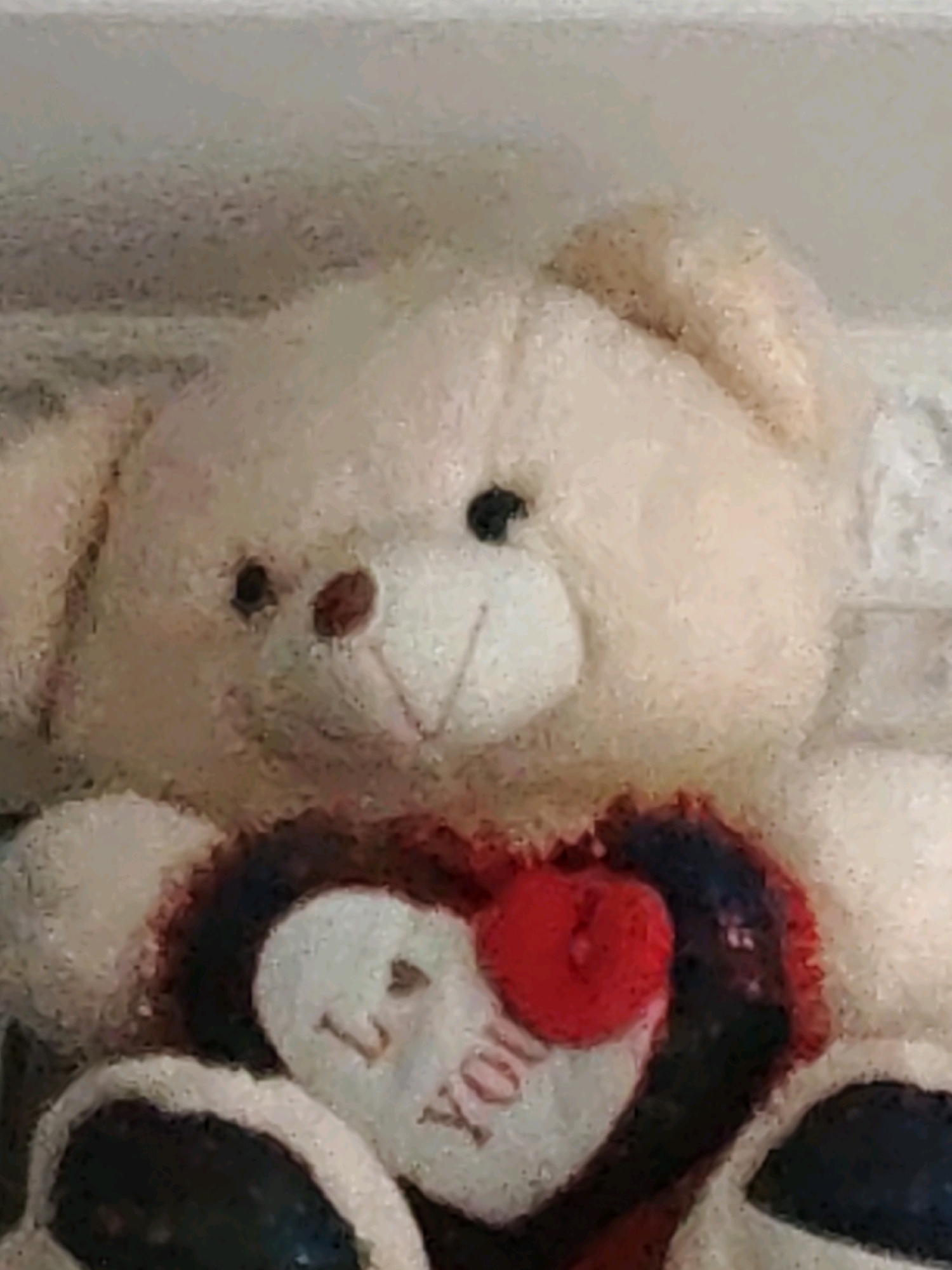 The image is a close-up, slightly blurry photograph of a cream-colored teddy bear. The bear is holding a heart-shaped pillow. The pillow is dark red with black accents and has the words "I LOVE YOU" written on it in white. The teddy bear has black eyes and a brown nose and mouth. The background is out of focus and appears to be a light-colored surface. The photograph is taken from a low angle, looking up at the teddy bear. There is no indication of the location in Ninotsminda, Georgia.