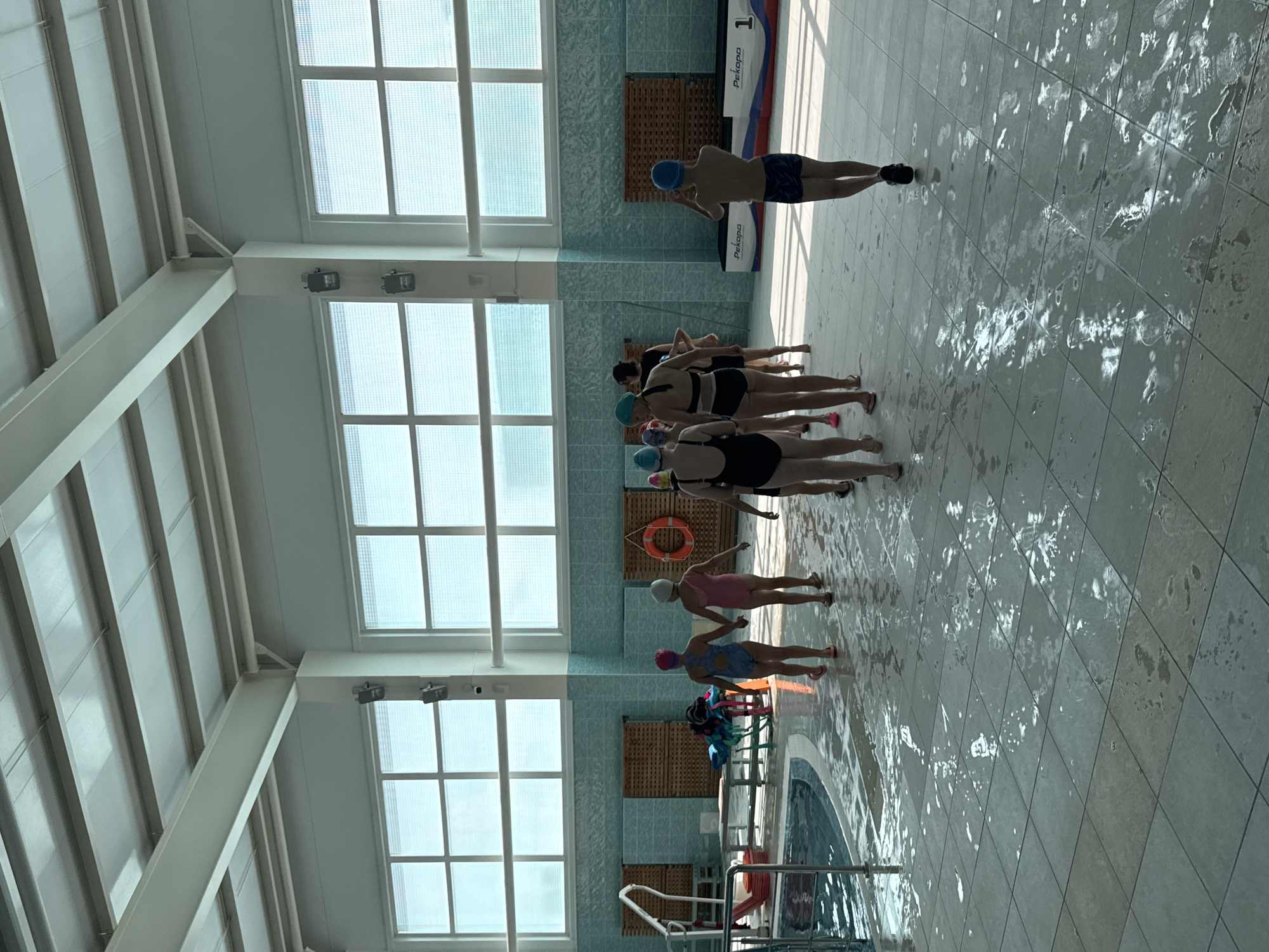 A group of children, some wearing swim caps and swimsuits, walk on a wet tiled floor in a well-lit indoor swimming pool facility. The pool is visible on the left side of the image. The walls are tiled with light blue ceramic tiles. Large windows with gridded panes are situated high on the walls, allowing natural light to enter. In the background, there are wooden lockers and a life preserver hanging on the wall. Two podiums, one labeled "Pekopa" and "1", are visible on the right. The floor is slick with water, suggesting recent activity in the pool area.