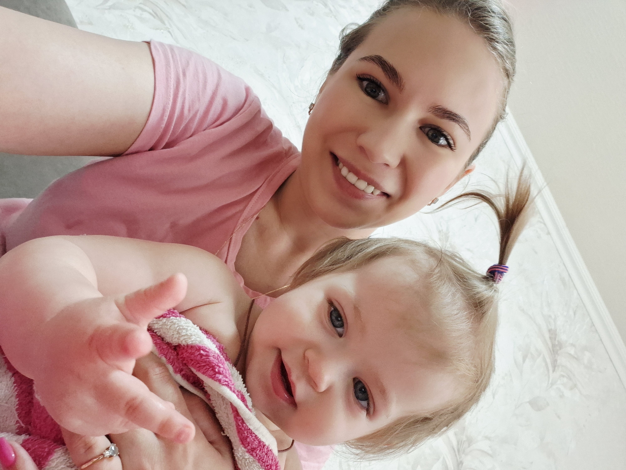 A selfie of a woman and baby, possibly taken indoors. The woman, with fair skin and light brown hair pulled back in a ponytail, is wearing a pink t-shirt and smiling. She has visible eye makeup and is wearing earrings and a ring. The baby, also fair-skinned with blonde hair in a small ponytail, is wrapped in a pink and white striped towel and reaching towards the camera with one hand. The background appears to be a patterned wall. There are no visible indications that this photo was taken in Chaikovsky, Russia.