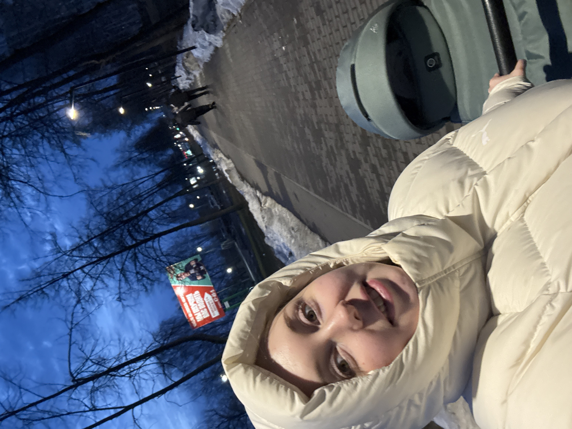 The image depicts an outdoor winter scene at dusk or night, located in Unknown, Unknown, on a paved pathway lined with bare trees and dirty snow. In the foreground, a light-skinned female, approximately in her twenties, with light hair and eyes, wears a cream-colored hooded puffer jacket featuring a visible Puma logo on the left sleeve. She is smiling directly at the camera, holding the black handle of a dark green baby stroller, branded "Aire," which is partially visible on the right. In the middle ground, two indistinct figures in dark clothing walk away from the camera on the wet pathway. Several streetlights illuminate the scene. In the background, a red and green billboard displays Russian text translating to "WE WILL TEACH YOU TO DRIVE," alongside an image of a man and a phone number "47-45-5." The sky above is a deep blue, partly cloudy, and distant urban lights are visible further down the path.