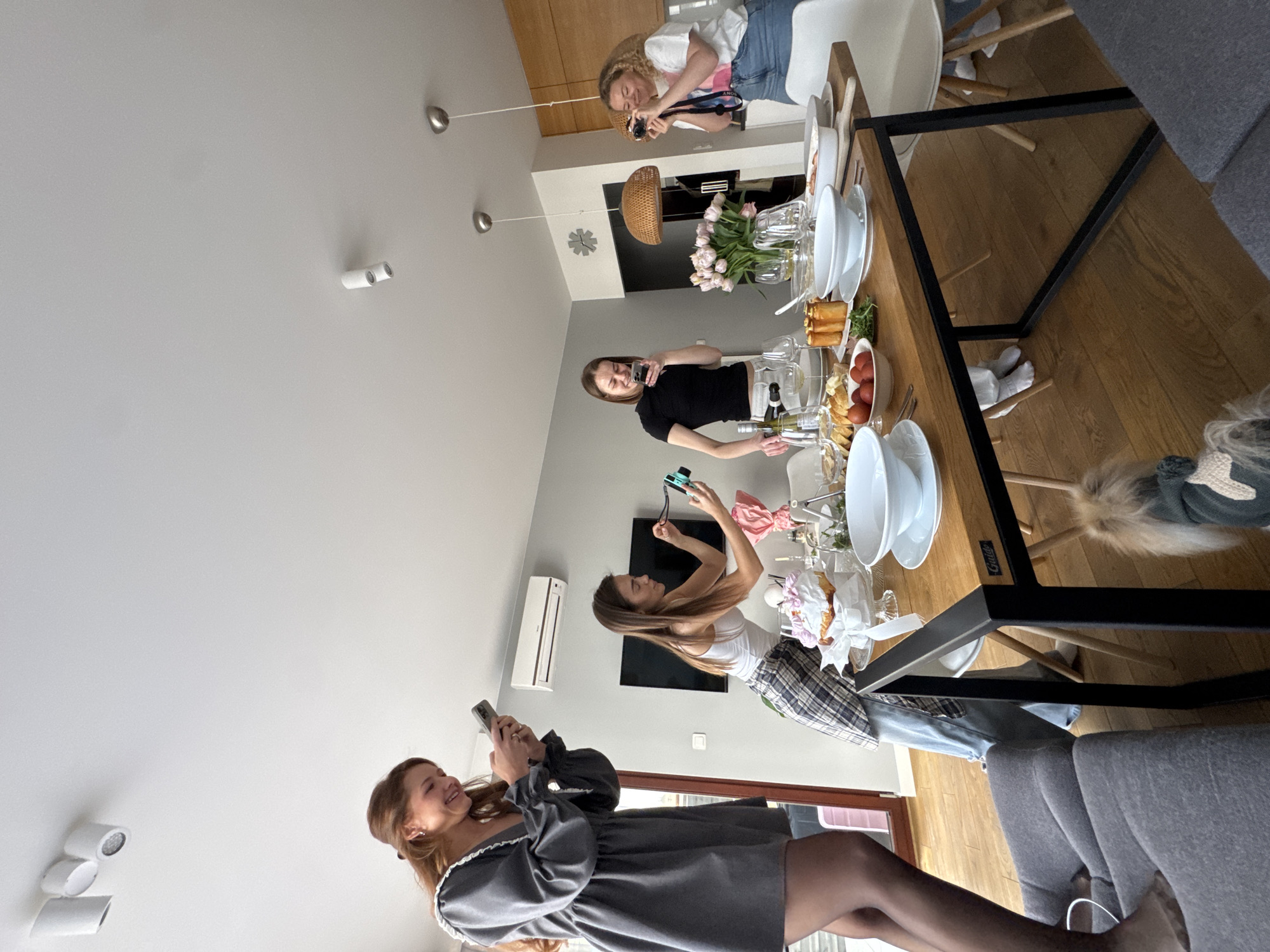 The image depicts an indoor scene with a group of four young women gathered around a table. They all appear to be taking pictures of the table's contents with their mobile phones or cameras. The table is set with plates, glassware, flowers, and various food items.
The room is brightly lit and features a minimalist design with white walls and light-colored wooden floors. There is a television on the wall.
Based on the style of the interior and the clothing of the people, the location is likely a modern apartment in Warsaw, Poland. There is no concrete evidence to confirm this hypothesis, but the style is consistent with modern Polish designs.