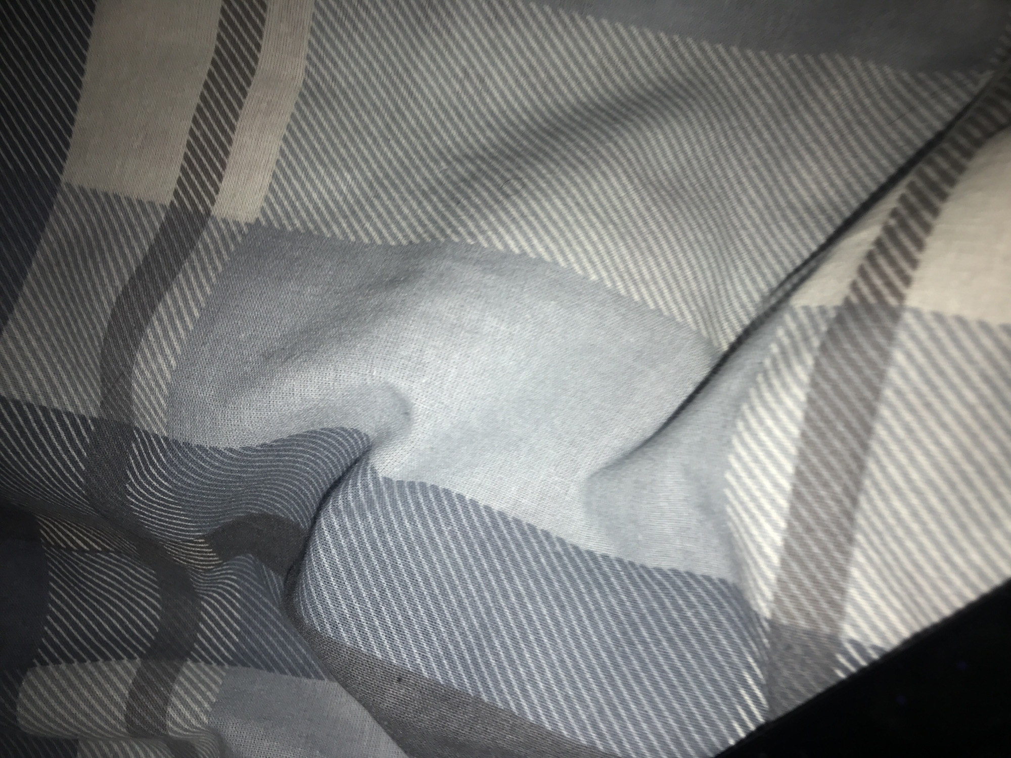 A close-up photograph depicts a folded fabric with a blue and gray plaid pattern. The material appears soft and textured, with visible seams and creases. The lighting casts shadows, accentuating the fabric's folds and the woven texture. The location is not discernible from the image.