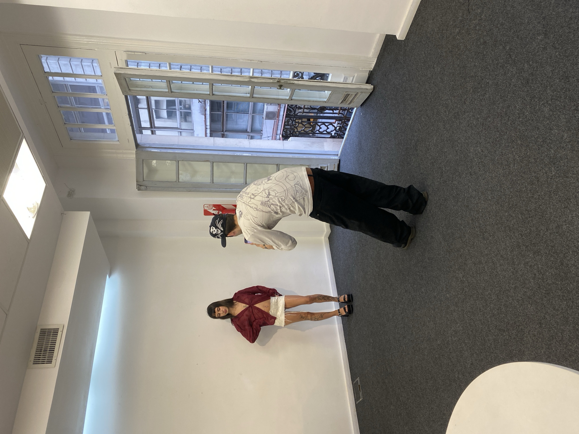 The image, correctly oriented by rotating it 90 degrees clockwise, depicts an indoor room in Buenos Aires, Argentina, featuring two individuals. The room has stark white walls and a dark grey, flecked carpet.
On the right side of the frame, a man, mostly seen from behind, is positioned near an open set of multi-paned white wooden double doors. He wears a white long-sleeved shirt with black patterns, black pants, and a black baseball cap. He appears to be leaning or looking out through the doors, which open onto an ornate wrought-iron balcony. Beyond the balcony, other urban buildings with windows are visible under bright daylight. A red object, possibly a fire extinguisher, is partially visible on the wall near him.
On the left, a woman stands facing generally towards the viewer, leaning against the white wall. She is dressed in a maroon or dark red sheer long-sleeved top, a white mini skirt, and black sandals. Her legs are adorned with distinct white or light-colored abstract tattoos. She has dark hair.
The room is illuminated by square recessed lights in the ceiling and a vertical strip light along a wall segment. A white rectangular air vent is also present on a wall. A partial view of a white, round table or stool is visible in the bottom right corner of the image. The setting combines contemporary interior elements with the classic architecture visible through the windows, suggesting a bright, open space. No discernible text is clearly visible on any objects or clothing.