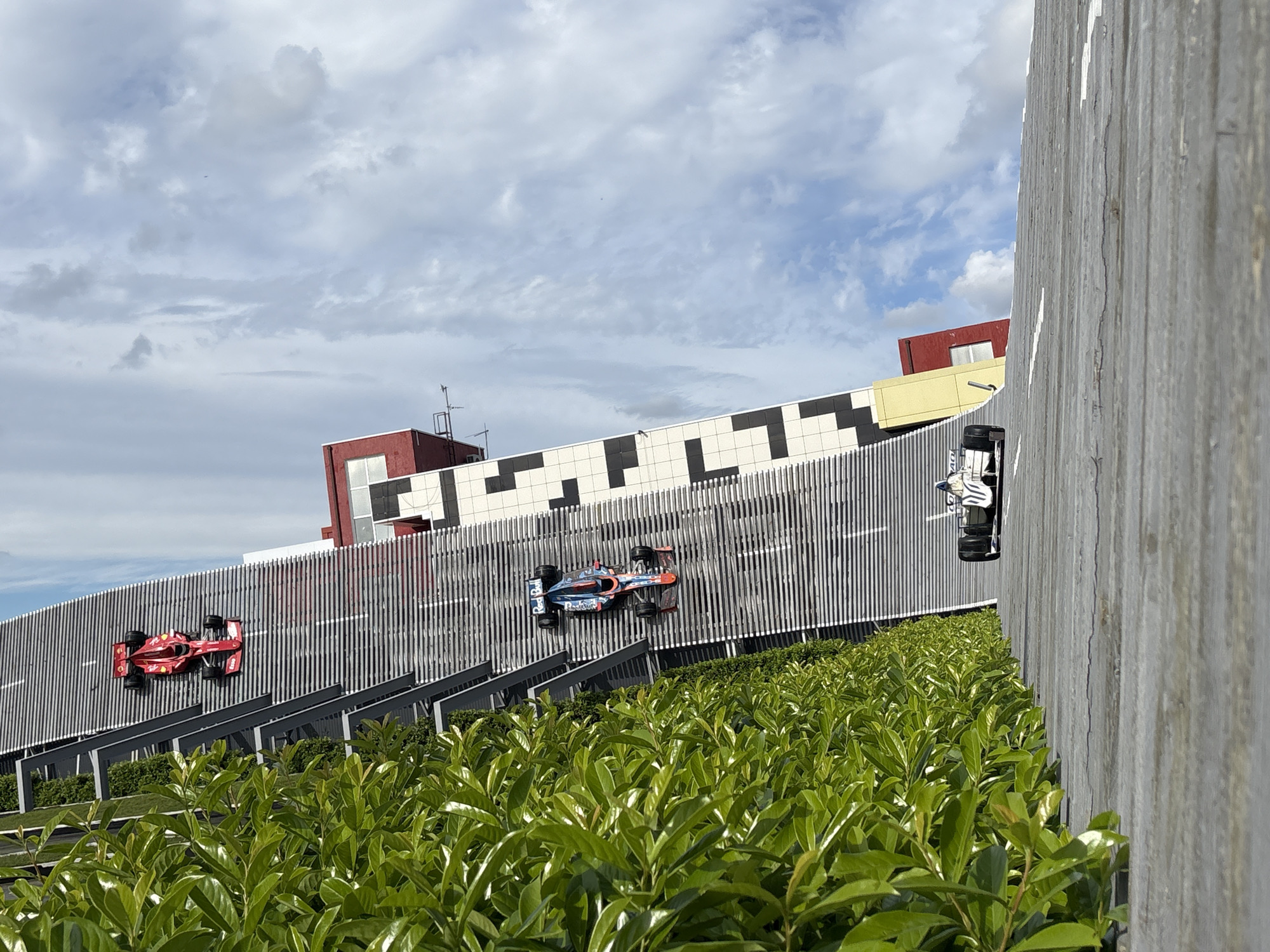 The image shows a street scene in Anapa, Russia, featuring a decorative representation of a race track with model Formula 1 cars on the side of a building. There are three Formula 1 cars visible: a red Ferrari, a blue-and-red Red Bull, and a white-and-black car. The decorative track is constructed with metal bars over a green shrub hedge. In the background is a building with a white, black, red and yellow facade. The sky is cloudy. A gray wooden structure is visible in the foreground, partially obscuring the view. The image is angled as if taken from ground level with an obstructed view.