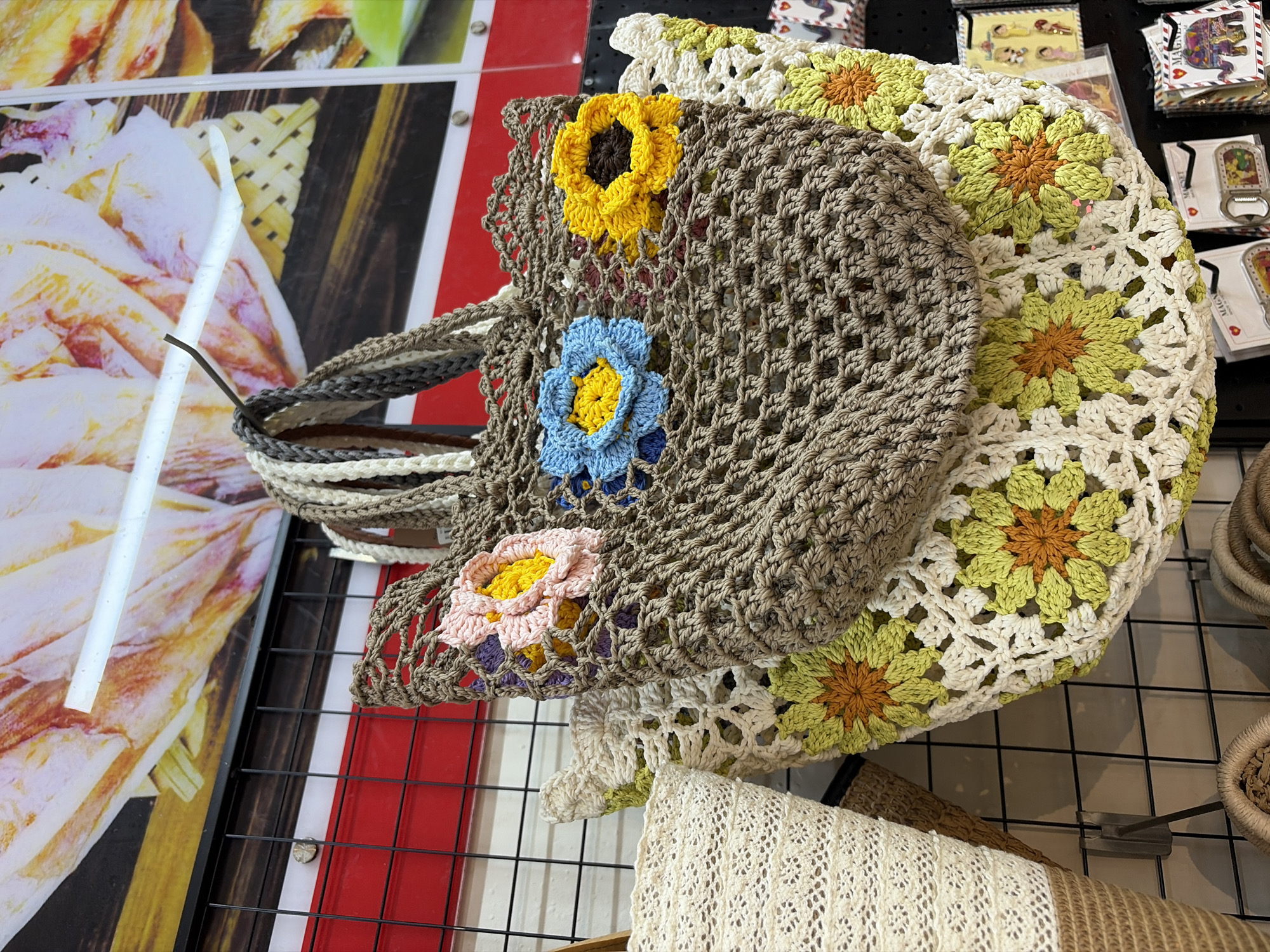 Two crocheted bags are displayed on a black wire grid in a shop. The bag in the foreground is large and white with a pattern of green and orange flowers. The bag behind it is smaller, made of brown crocheted material, and adorned with crocheted flowers in pink, blue, and yellow. A third, cream-colored bag is partially visible on the left. The background shows a colorful display of posters and items on shelves. The overall impression is of a vibrant, handcrafted textile display, likely in a market or boutique. This scene is located in Đà Nẵng, Vietnam.