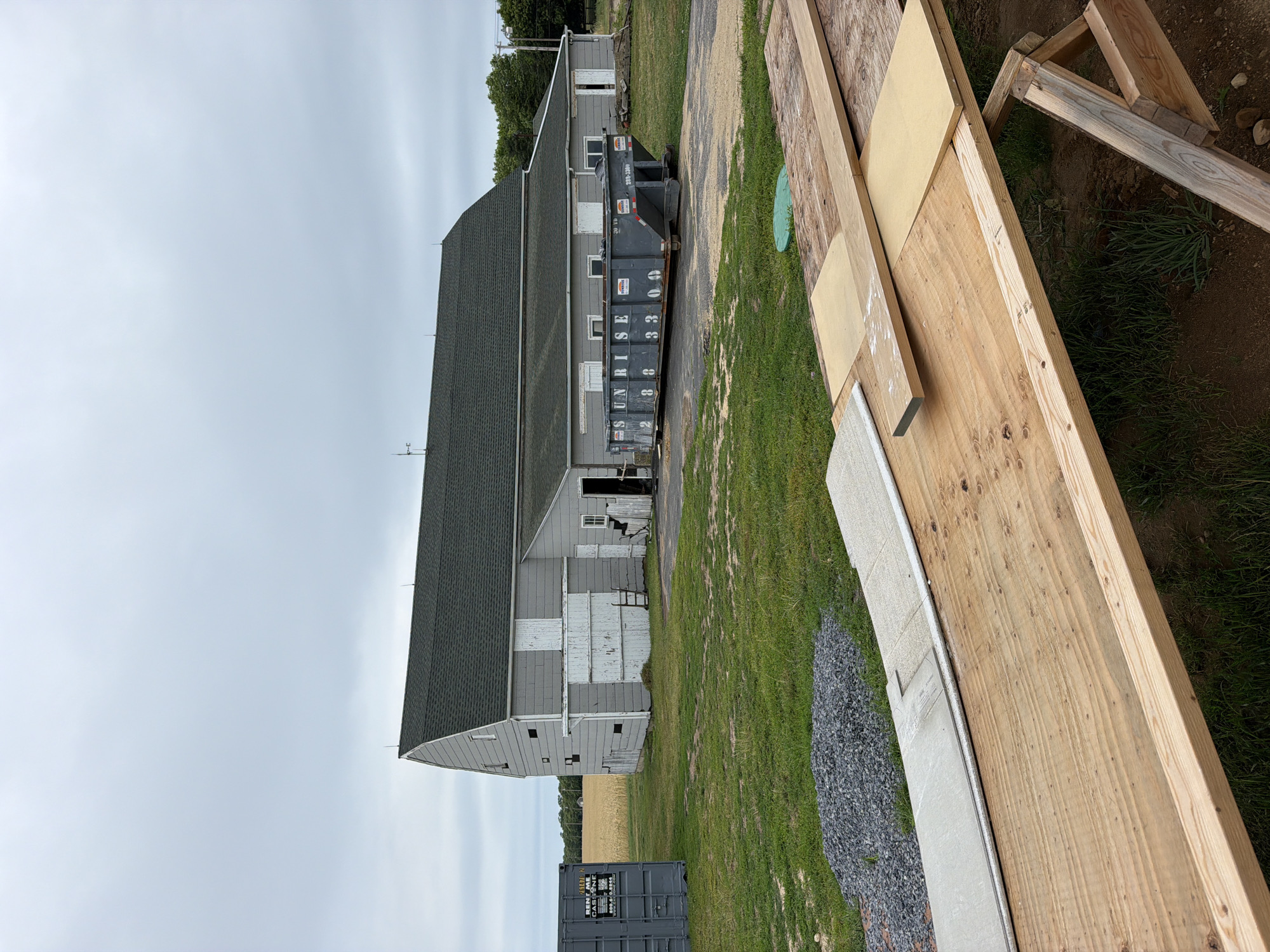 This image shows a construction site with a large, gray building in the background and wooden materials in the foreground. The building appears to be a barn or a similar agricultural structure, with a dark green roof and light gray siding. There are several windows and doors on the building, some of which are boarded up. A large, dark gray dumpster is parked next to the building, with "SUNRISE" written on its side in white letters, along with a phone number.
In the foreground, there are various construction materials, including large sheets of plywood and wooden beams. Some of the wood appears to be freshly cut, while other pieces show signs of wear. There is also a pile of gravel and a section of what looks like a concrete curb or foundation. The ground is covered in grass and dirt. The scene appears to be an outdoor construction project.
The sky is overcast, suggesting it is either early morning or late afternoon, or a cloudy day. There are no people visible in the image. The overall atmosphere is one of ongoing work or preparation for construction. The location is identified as Mattituck, United States of America.