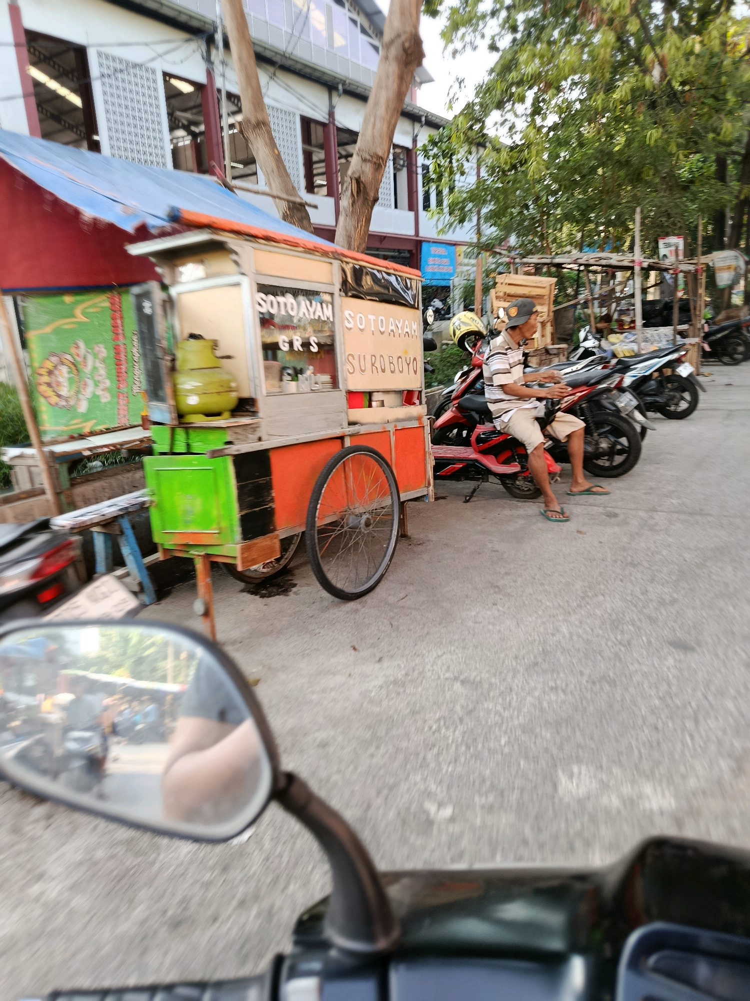 In Bekasi, Indonesia, a street vendor operates a food cart, likely selling "Soto Ayam" (chicken soup) as indicated by the signage. The cart, primarily orange and red with blue accents and a green panel, is equipped with a large green gas cylinder. A man, wearing a striped shirt, shorts, and flip-flops, is seated on a motorcycle parked next to the cart. Several other motorcycles are visible behind him, suggesting a common mode of transportation in the area. The scene unfolds on a paved street, with trees providing shade and a building with patterned windows in the background. The image is captured from a moving vehicle, as evidenced by the blurred foreground and the reflection in a motorcycle's rearview mirror.