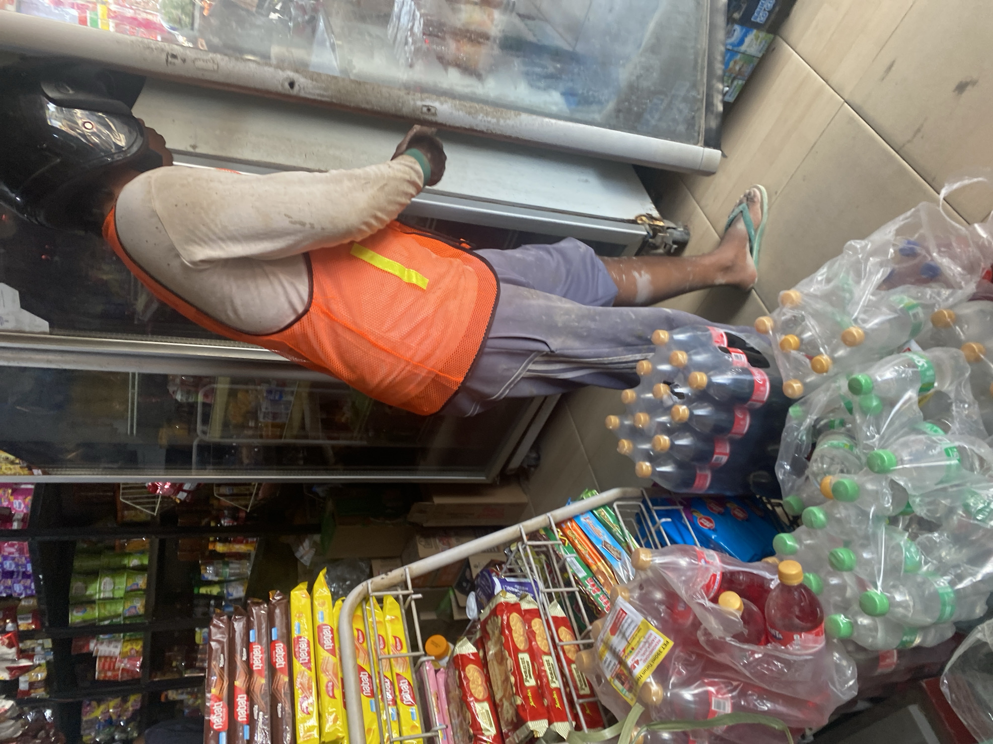 In a narrow aisle of a small convenience store in Bulukumba, Indonesia, a person wearing a dark helmet, a light-colored long-sleeved shirt, and an orange safety vest is reaching into a glass-fronted refrigerator. The person's lower body is clad in dusty grey shorts and a teal flip-flop on their right foot. The store's interior is densely packed with merchandise. To the left of the refrigerator, stacked on a metal rack, are numerous packaged snacks, including what appear to be Nabati wafers in yellow and brown packaging, and other boxed and bagged items. In the foreground, several large plastic bags contain rows of bottled beverages, predominantly clear bottles with green caps and some darker liquid-filled bottles with red labels. The floor is made of light-colored, wood-patterned tiles. The overall impression is one of a busy, possibly informal, retail environment.