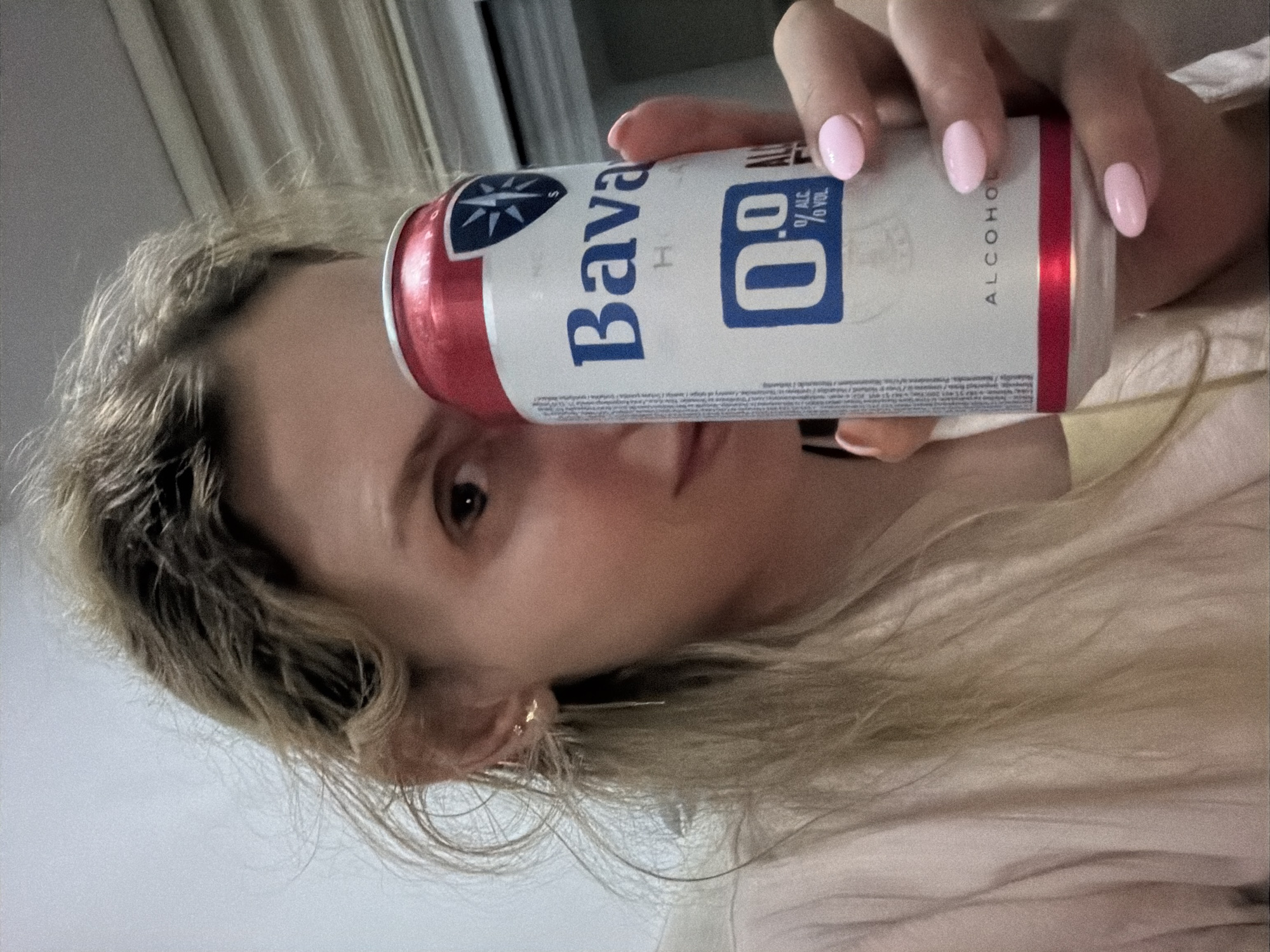 A woman with blonde hair and fair skin holds a can of "Bavaria 0.0" non-alcoholic beer in front of her face. Her nails are painted a light pink color. She is wearing a light pink shirt or top. The background is blurry, but appears to be a room with white curtains. The specific location in Budva, Montenegro, cannot be determined from this image.