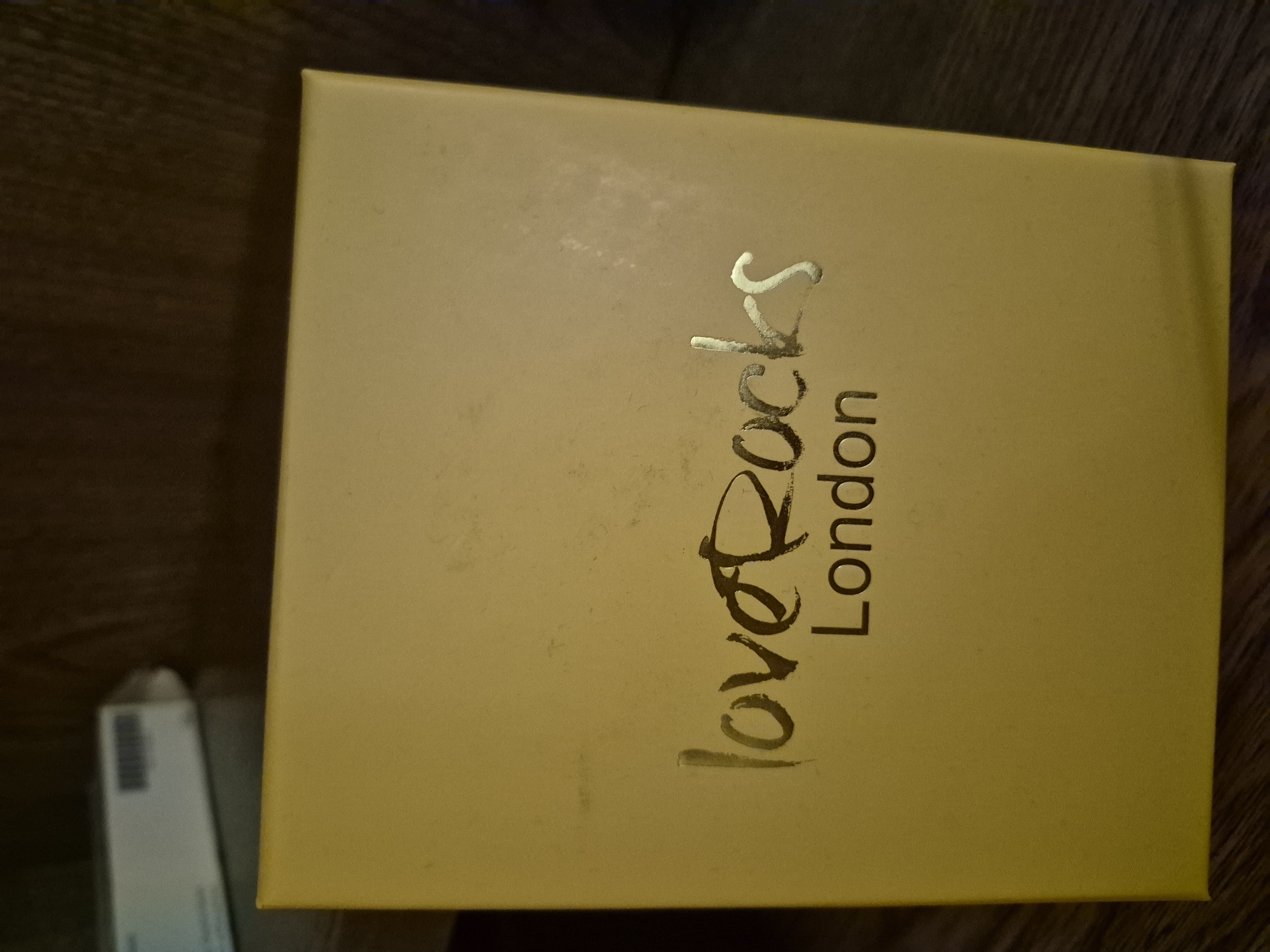 A close-up, eye-level shot shows a yellow box with the words "loveRoc/ London" printed in gold. The lettering is stylized and somewhat worn. The box is positioned on a dark wooden surface. In the upper left corner of the image, a blurry white rectangular object with a barcode is visible, suggesting it might be another product or packaging. The background is dark and out of focus, with the texture of what appears to be upholstery visible. The lighting is dim, casting soft shadows.