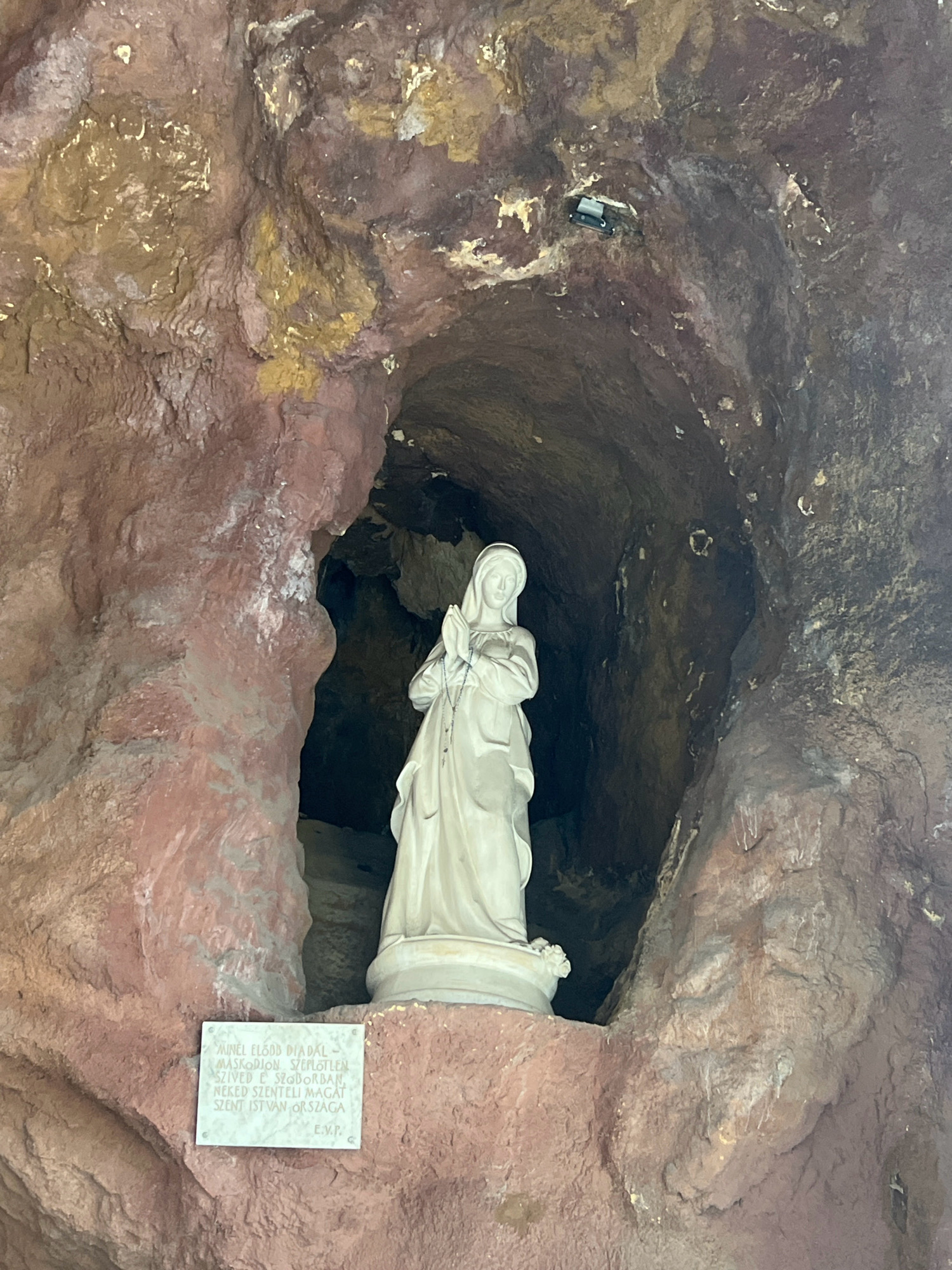 This is a full-shot, eye-level, indoor photograph of a statue of the Virgin Mary within a grotto. The statue is white and depicts Mary with her hands clasped in prayer. She is wearing a simple robe and veil. The grotto walls are rough and uneven, with reddish-brown and dark brown rock formations. There is a small, white plaque with text in front of the statue. The grotto is dimly lit, with some light illuminating the statue and the foreground. The scene is located in the Cave Church of Gellért Hill in Budapest, Hungary.