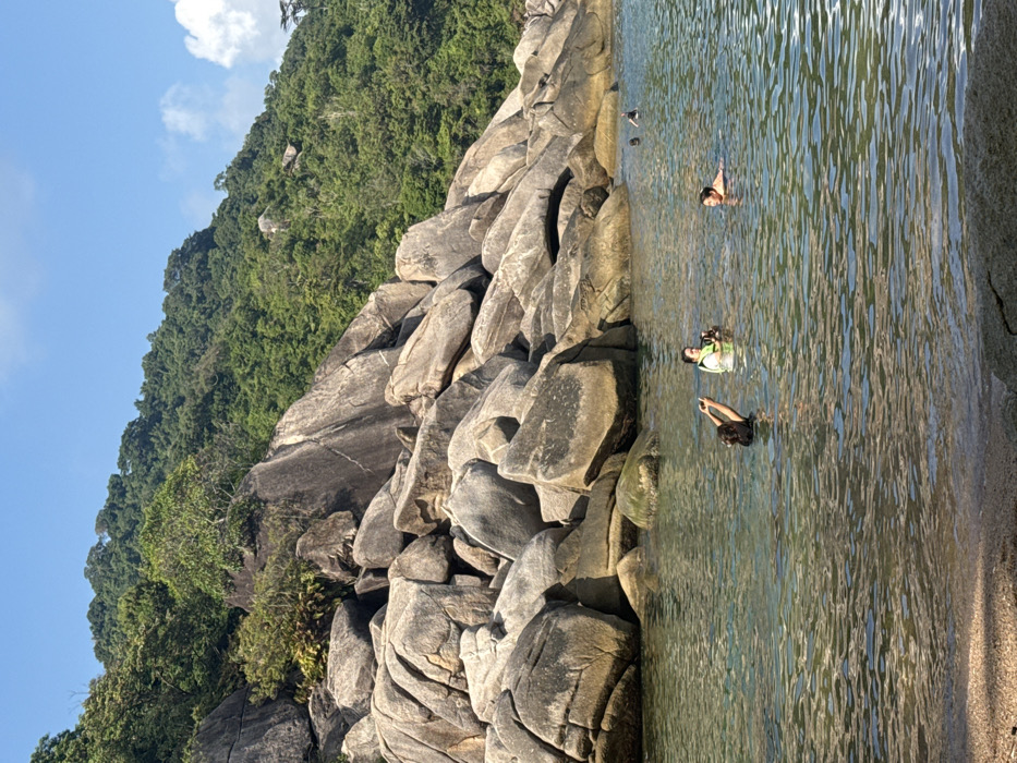 Unknown, Unknown: The scene captures a clear daytime view of a tropical coastal environment. A prominent formation of large, rounded grey boulders occupies the left and center mid-ground, extending into calm, clear turquoise water. Behind these rocks, a steep, densely vegetated green hill ascends, topped by a bright blue sky with sparse white clouds. Four individuals are observed in the water. In the foreground, a person in a dark top holds a smartphone above the water, oriented as if capturing an image. To their immediate left, a second individual wearing a light-colored life vest is partially submerged, holding a dark object. A third person is partially submerged to the right, facing the first two. Two more distant individuals are visible in the water in the far right background, one appearing to use a snorkel. The immediate foreground shows a shallow, sandy water's edge.