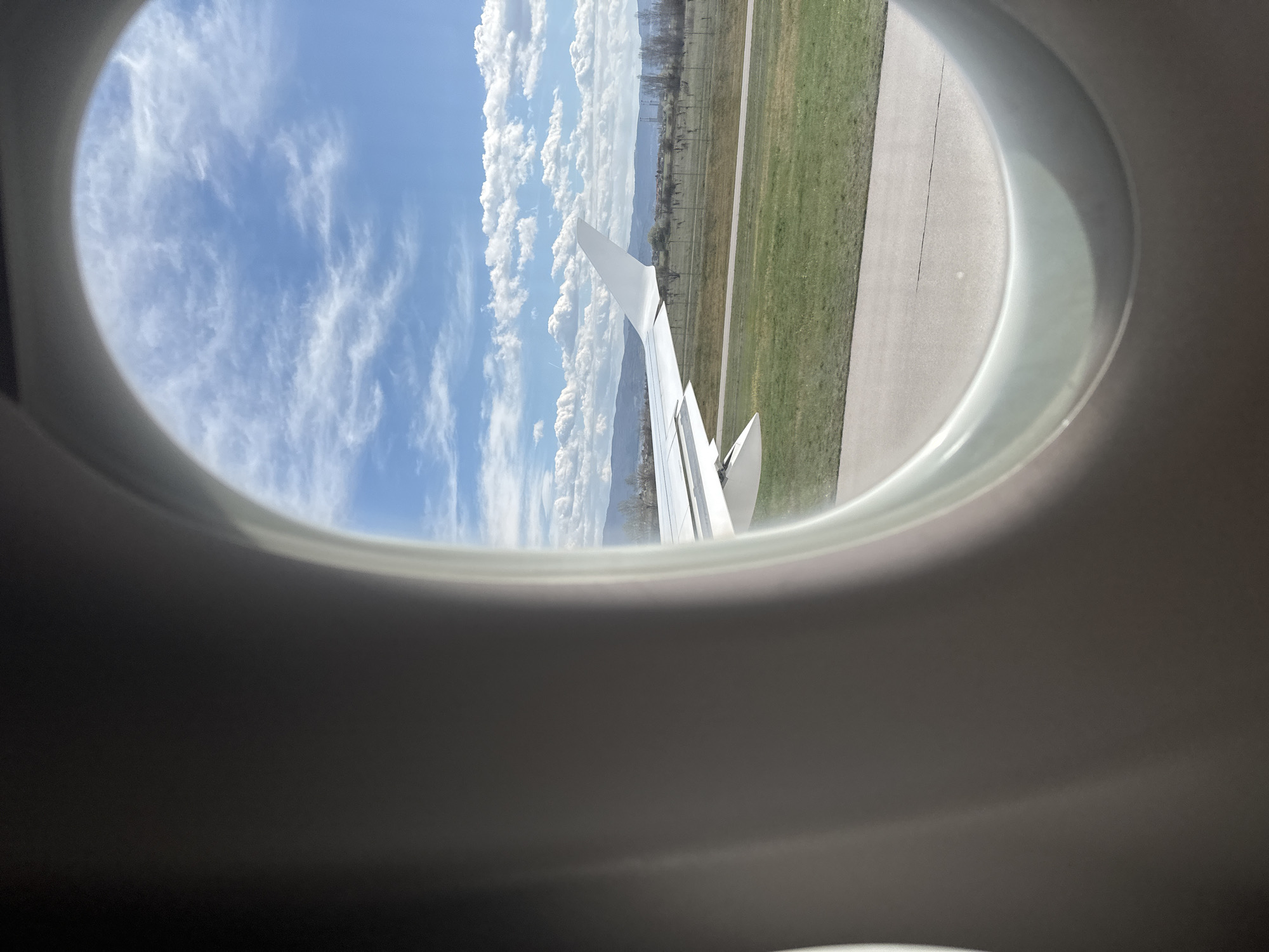 The image depicts a view from an airplane window. Visible outside is the plane's wing, a grassy area, a runway, some distant trees and buildings, a mountain range, and a partly cloudy blue sky. The photograph was taken from an aircraft during takeoff from Sofia Airport, Sofia, Bulgaria.
