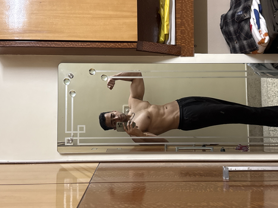 The scene is an indoor room, located in Kolkata, India. A shirtless adult male subject is centrally positioned, reflected in a large rectangular mirror. He is wearing black athletic trousers, a silver-colored chain around his neck, and a gold-colored bracelet on his left wrist. The subject holds a light-colored smartphone in his left hand, pointing it towards the mirror, and is depicted flexing his right bicep. His head is slightly tilted, and his gaze appears directed towards the smartphone screen.
The mirror has a decorative frosted glass border with geometric patterns and multiple circular glass or acrylic embellishments along its top and side edges. The immediate environment reflected in and surrounding the mirror consists of light-colored walls. To the viewer's left, a light-brown wooden wardrobe with a visible metallic handle is partially visible. To the viewer's right, a wooden cabinet structure features an open shelf containing a yellow item and several white papers. Below this open shelf, a stack of folded garments is visible, with a plaid shirt positioned on top. The flooring is a light, neutral color.