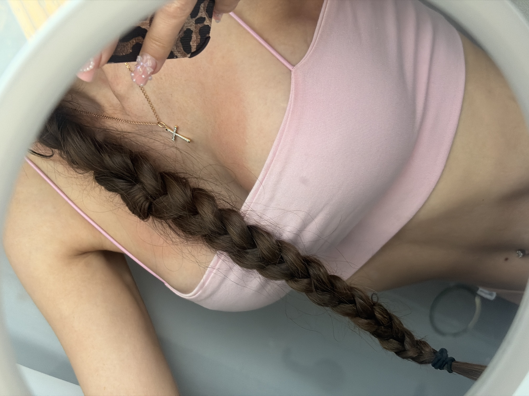 The image is a close-up, self-taken photograph, likely in a bathroom, showing a person's upper body from the chest up. The person is wearing a light pink ribbed tank top with thin spaghetti straps. A thick, dark brown braid of hair is visible hanging over their left shoulder and down their chest. Around their neck, they wear a delicate gold chain with a small, jeweled cross pendant. On their right side, a hand with long, artificial nails, decorated with pearls and floral designs, is visible near their neck, holding what appears to be a phone with a leopard print case. There are distinct red marks on the person's chest and neck, indicating a tan line or sunburn. The background is out of focus but appears to be a light blue wall or surface, with reflections in what looks like a mirror. The image does not provide any specific details or context related to Saint Petersburg, Russia.