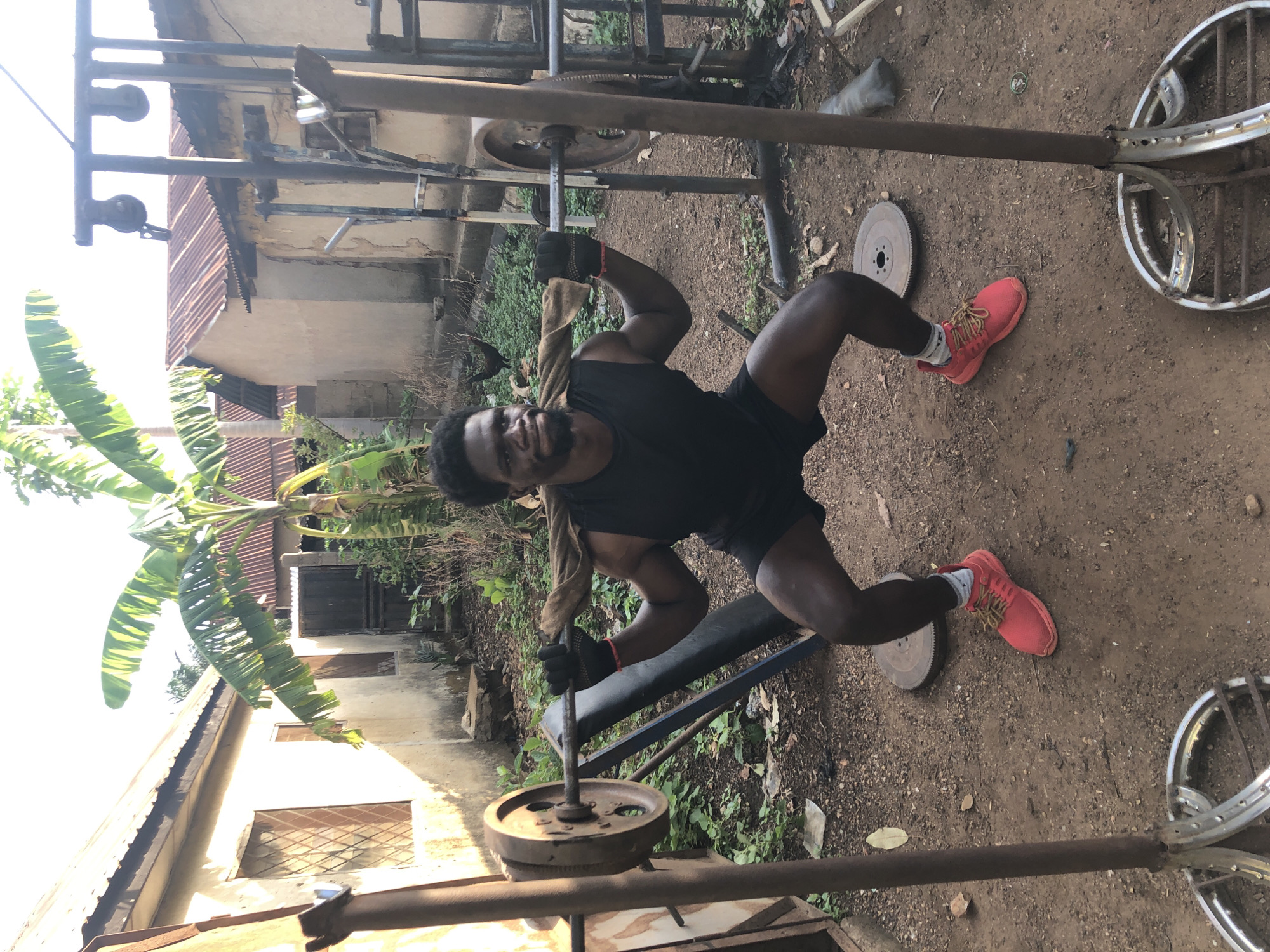 A man is exercising outdoors in Akwanga, Nigeria. He is wearing a black tank top, black shorts, red sneakers, and workout gloves. He is using a homemade weight bench press with repurposed metal weights and car rims. The environment is a dirt yard with scattered vegetation and several buildings in the background. There is a banana tree to the left. The exercise equipment is makeshift.