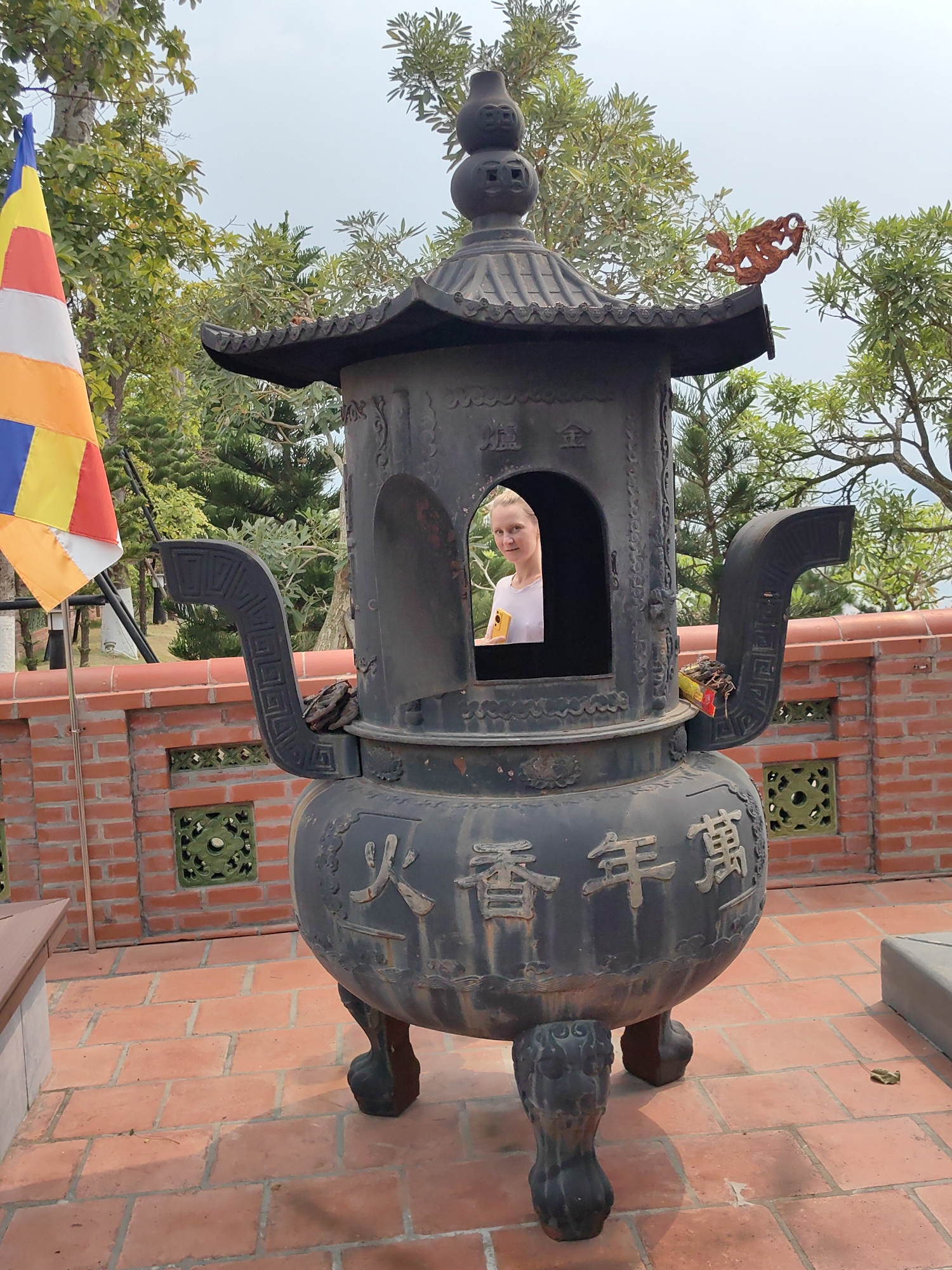 A woman stands inside a large incense burner at a temple in Hong Gai Ward, Vietnam. The burner, made of dark metal, has an ornate roof and handles, with Chinese characters inscribed on its side. A Buddhist flag stands to the left of the burner, and a low brick wall is visible behind it. The ground is paved with red bricks, and trees are visible in the background. The sky is overcast.